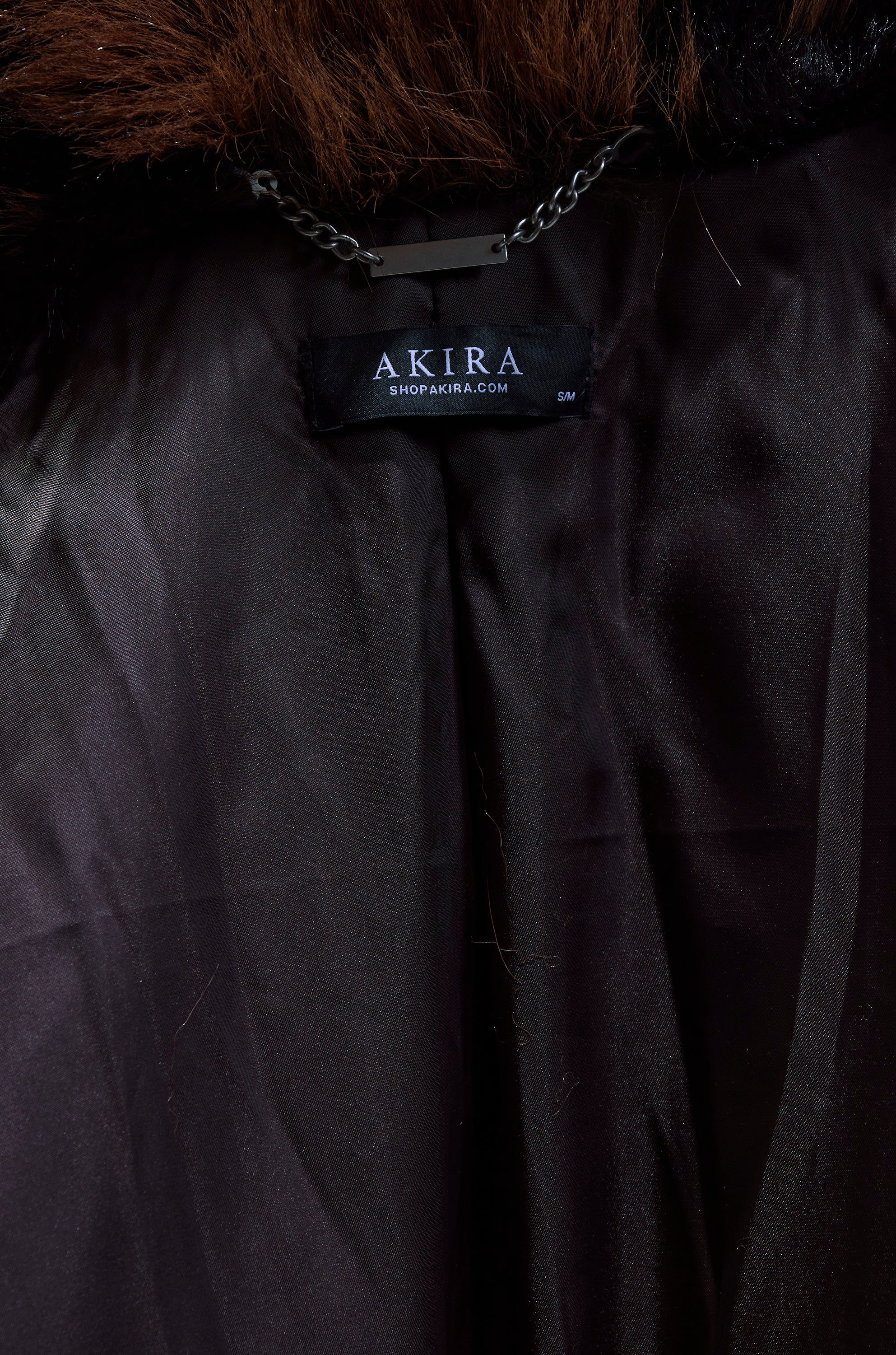 A close-up of the TELLURIDE FAUX FUR IN BROWN shows its black satin lining, an "AKIRA SHOPAKIRA.COM S/M" label, plush brown faux fur with an oversized collar, and a chain for hanging.