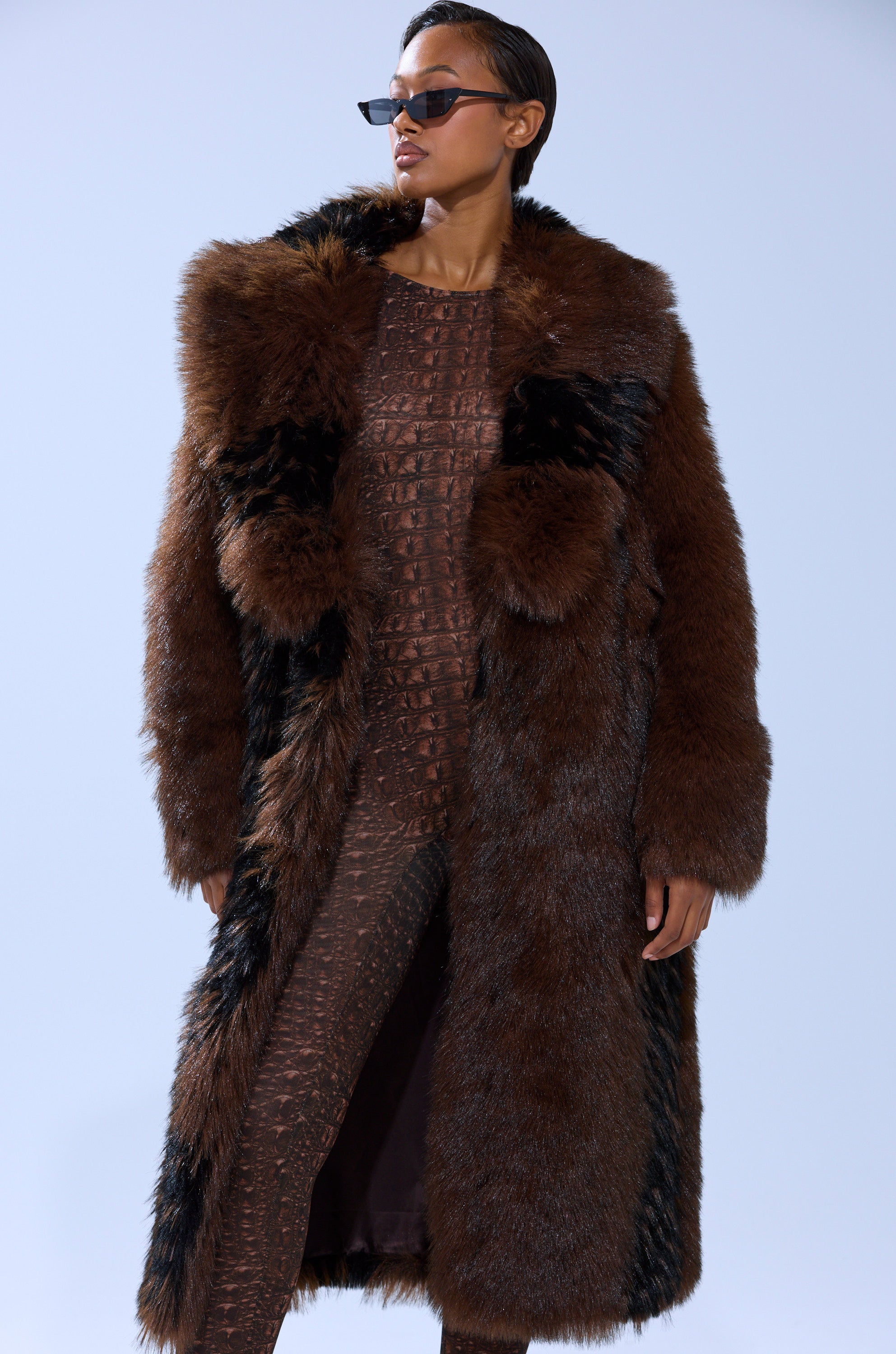 Wearing the TELLURIDE FAUX FUR IN BROWN jacket with black accents over a textured brown outfit and dark sunglasses, a person stands against a plain light background.
