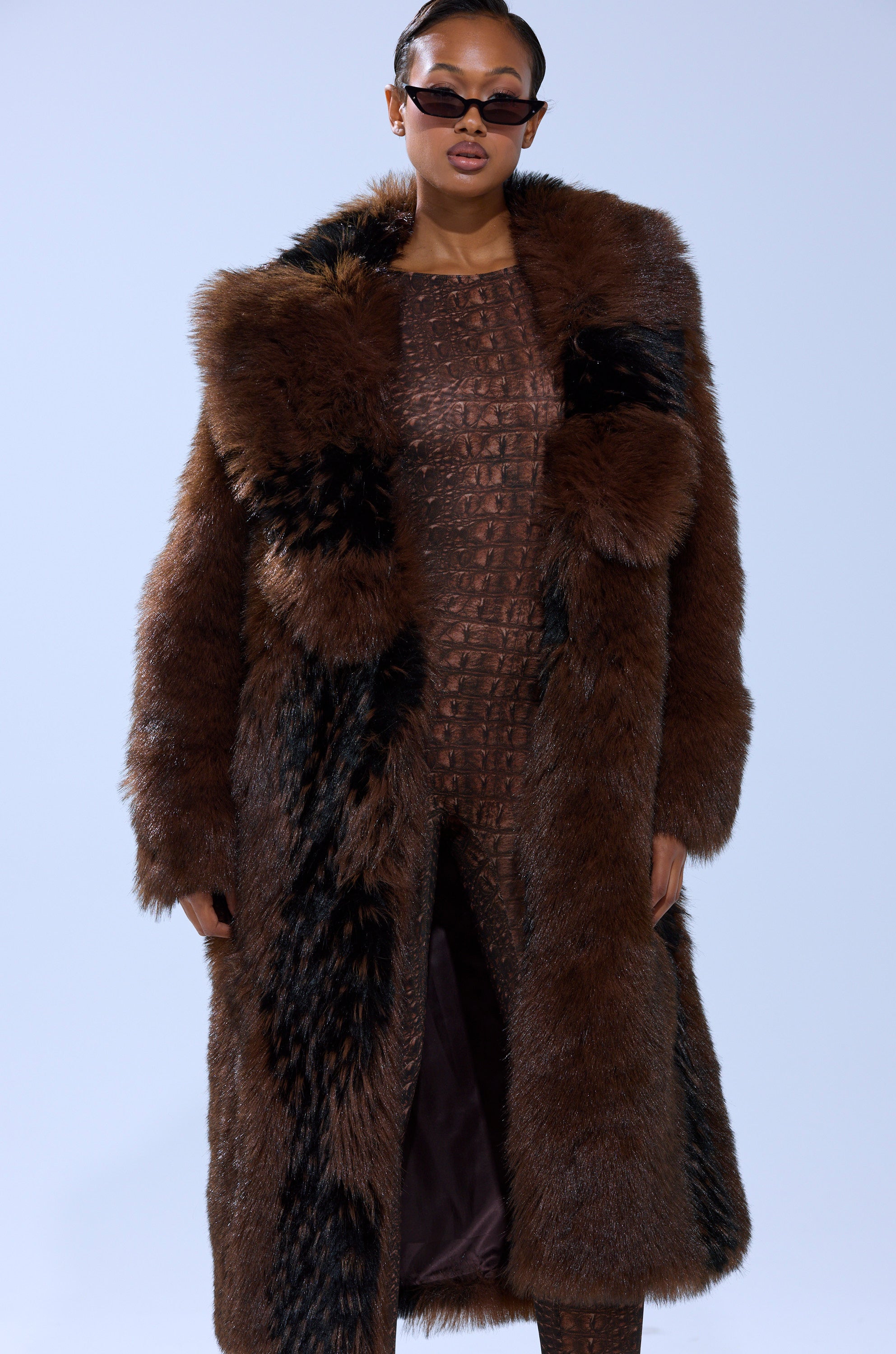 A woman in the TELLURIDE FAUX FUR IN BROWN jacket, dark sunglasses, and a textured brown outfit poses confidently against a plain light background.