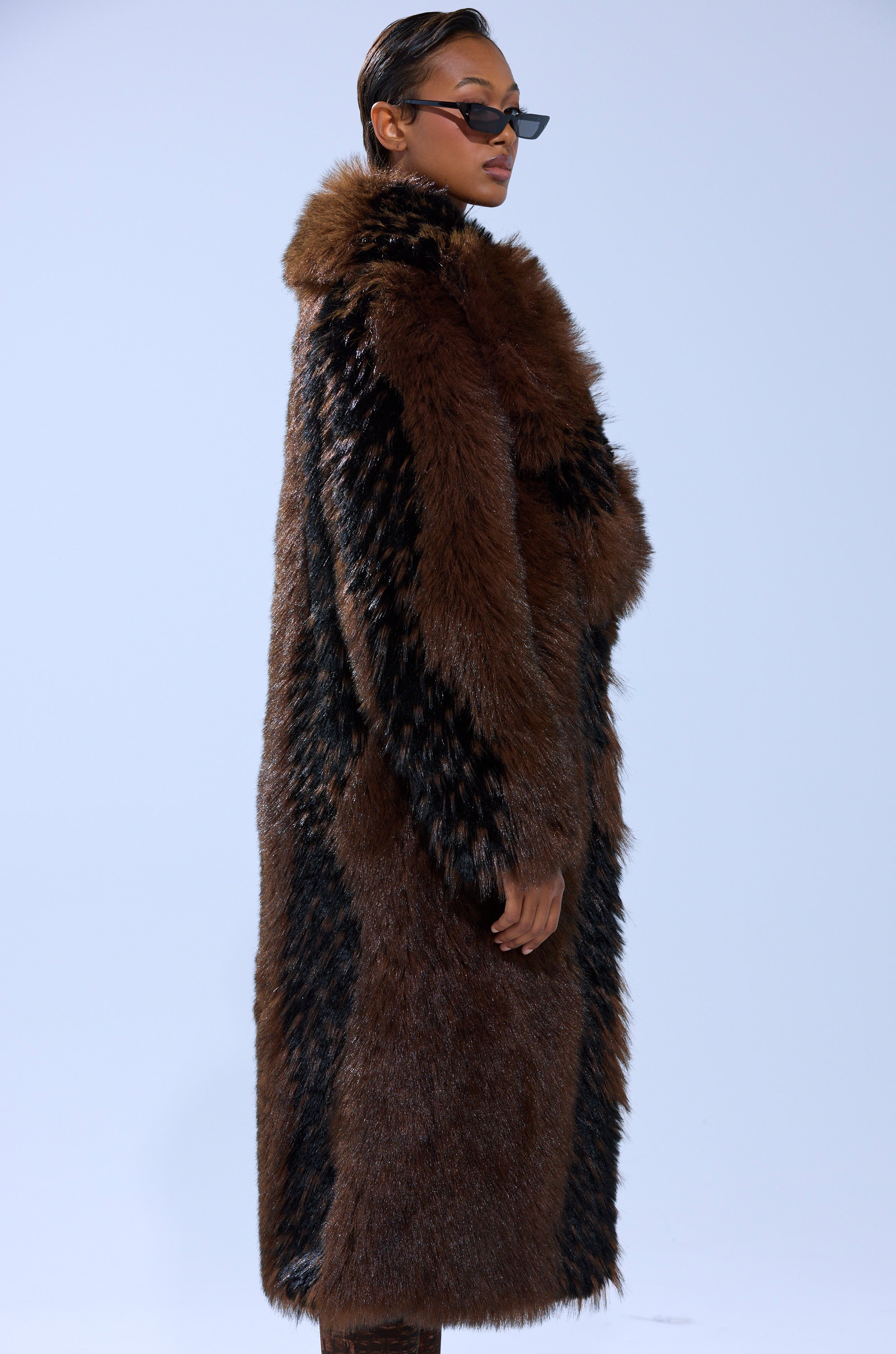 A person in dark sunglasses and the TELLURIDE FAUX FUR IN BROWN, featuring a luxurious longline silhouette with an oversized fit, stands sideways against a plain light background, exuding confidence.