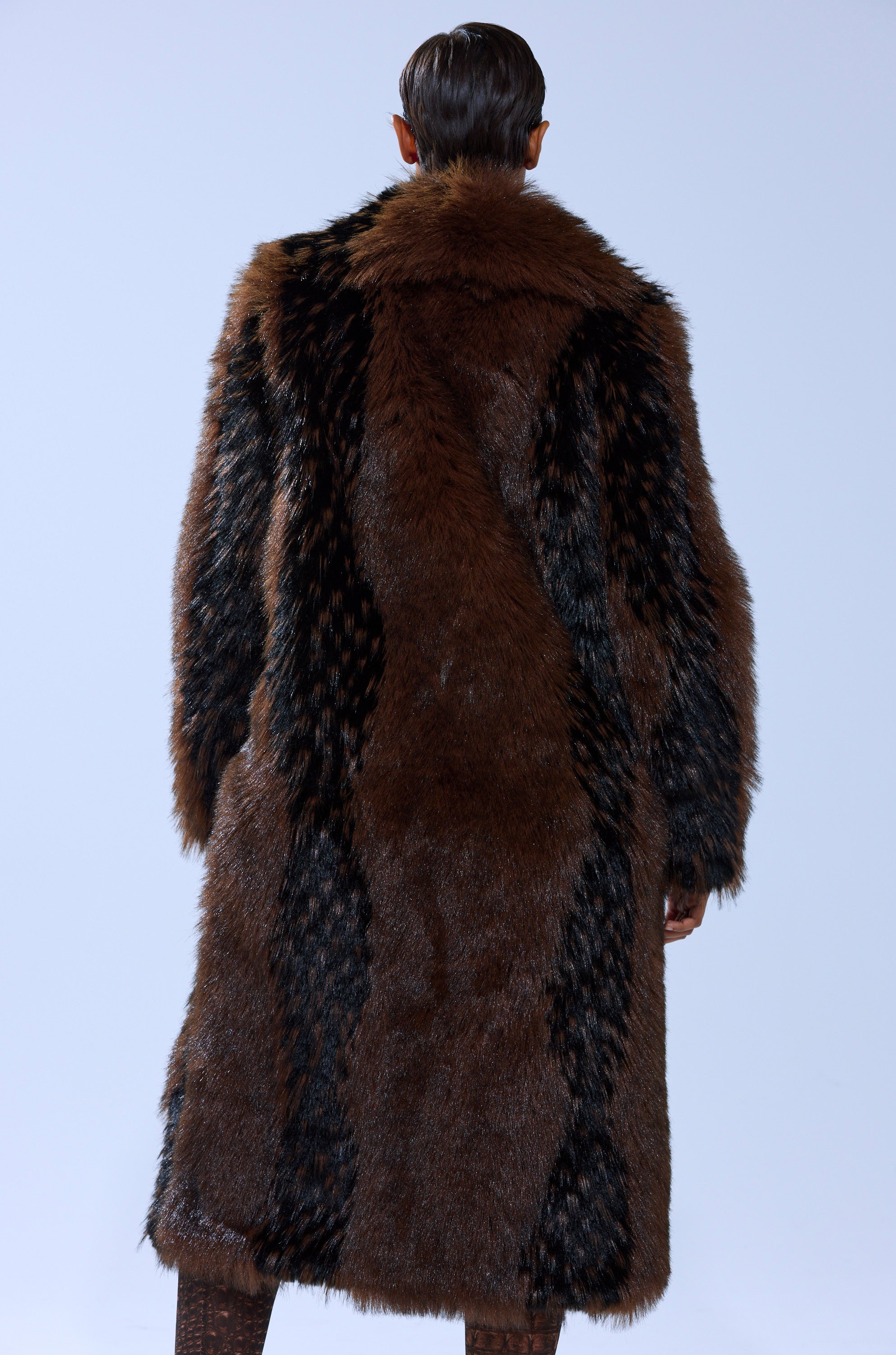 A person with short, dark hair is seen from behind, wearing the TELLURIDE FAUX FUR IN BROWN—an oversized, luxurious brown and black faux fur jacket that falls below the knees. The background is plain and light-colored.