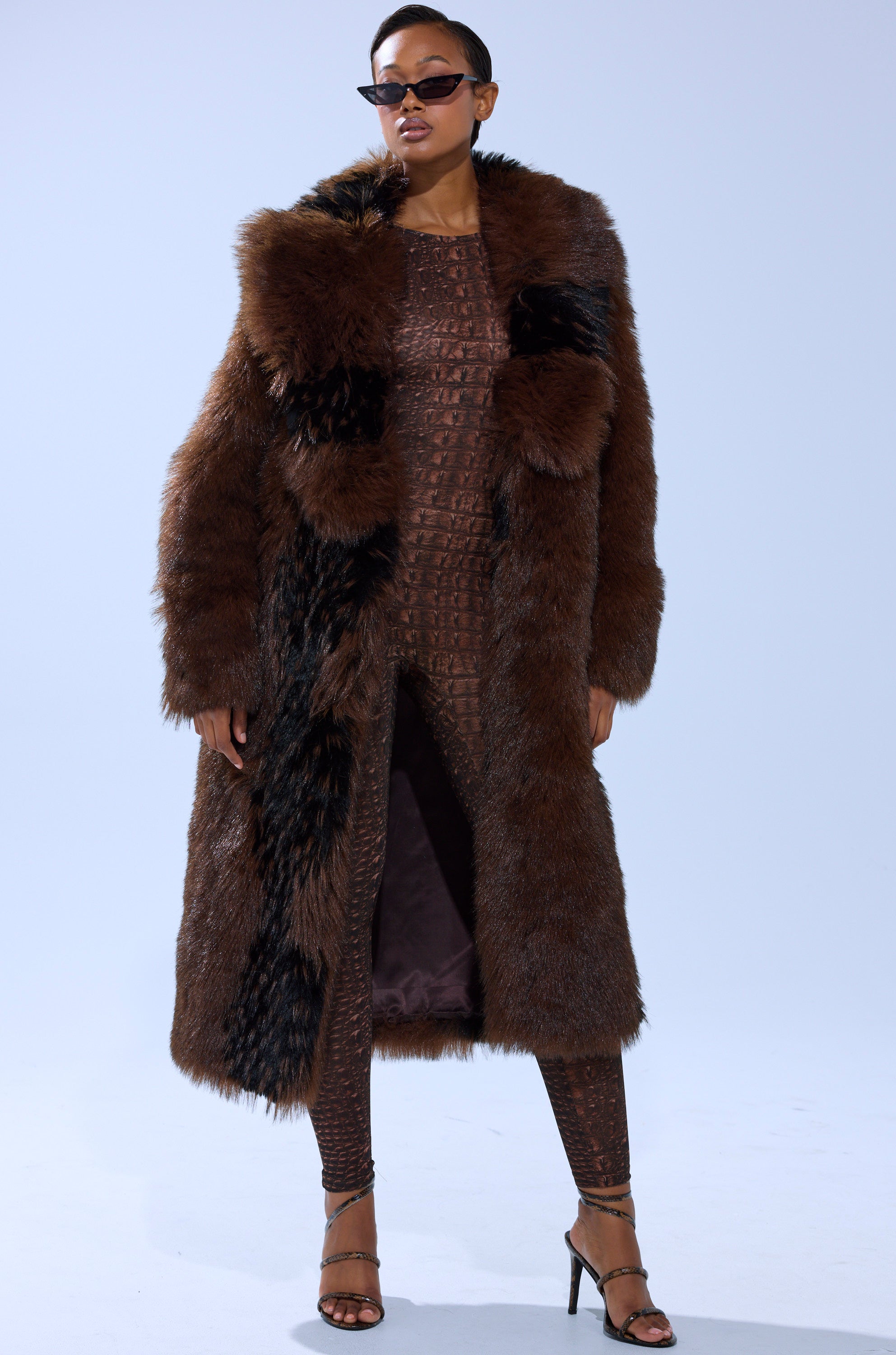 Wearing the TELLURIDE FAUX FUR IN BROWN over a brown textured bodysuit, with black sunglasses and strappy high-heeled sandals, a person stands against a plain light background.