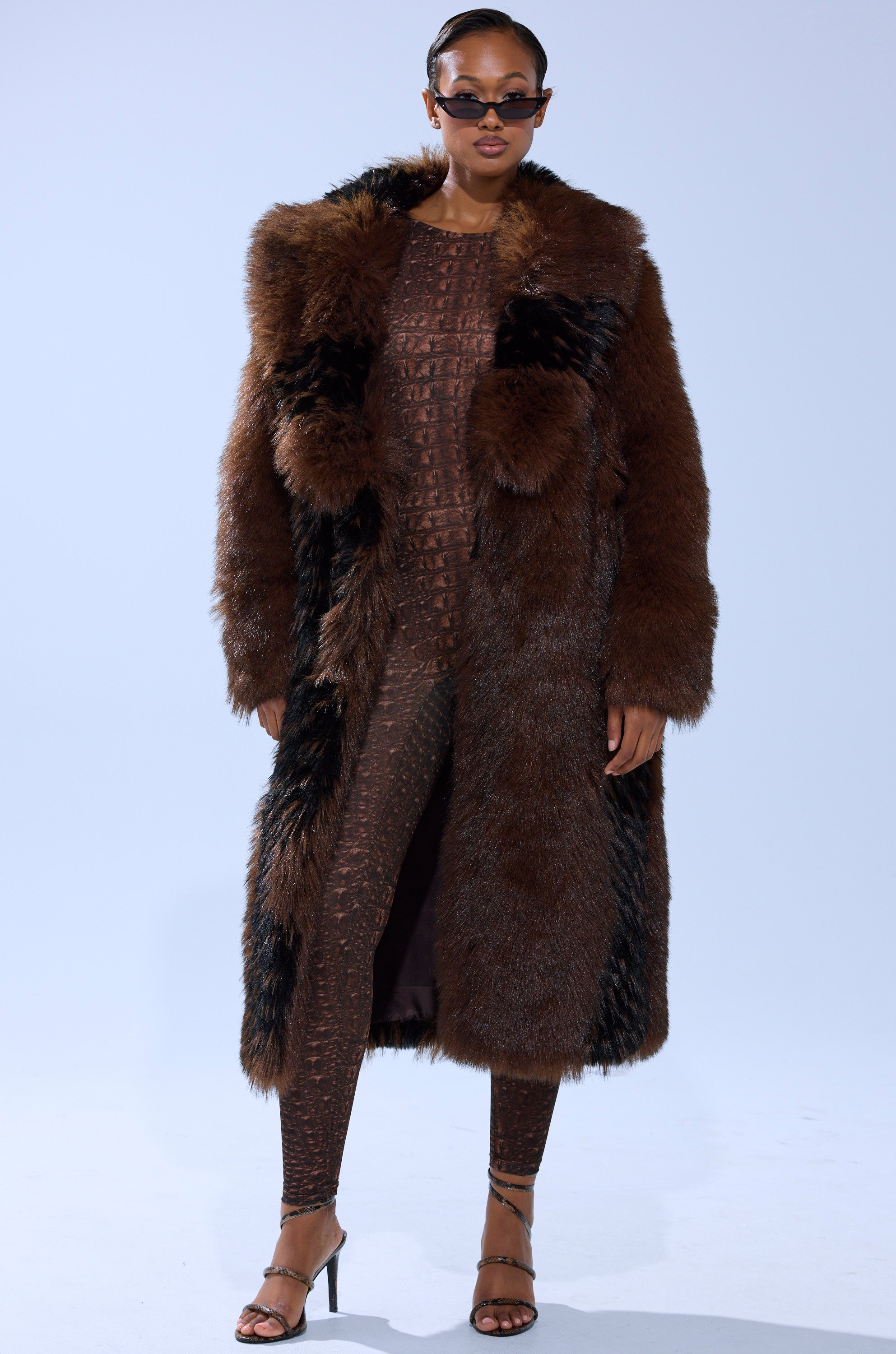 Wearing sunglasses, the model stands confidently against a plain light background in the TELLURIDE FAUX FUR IN BROWN layered over a textured brown outfit and open-toe heels.