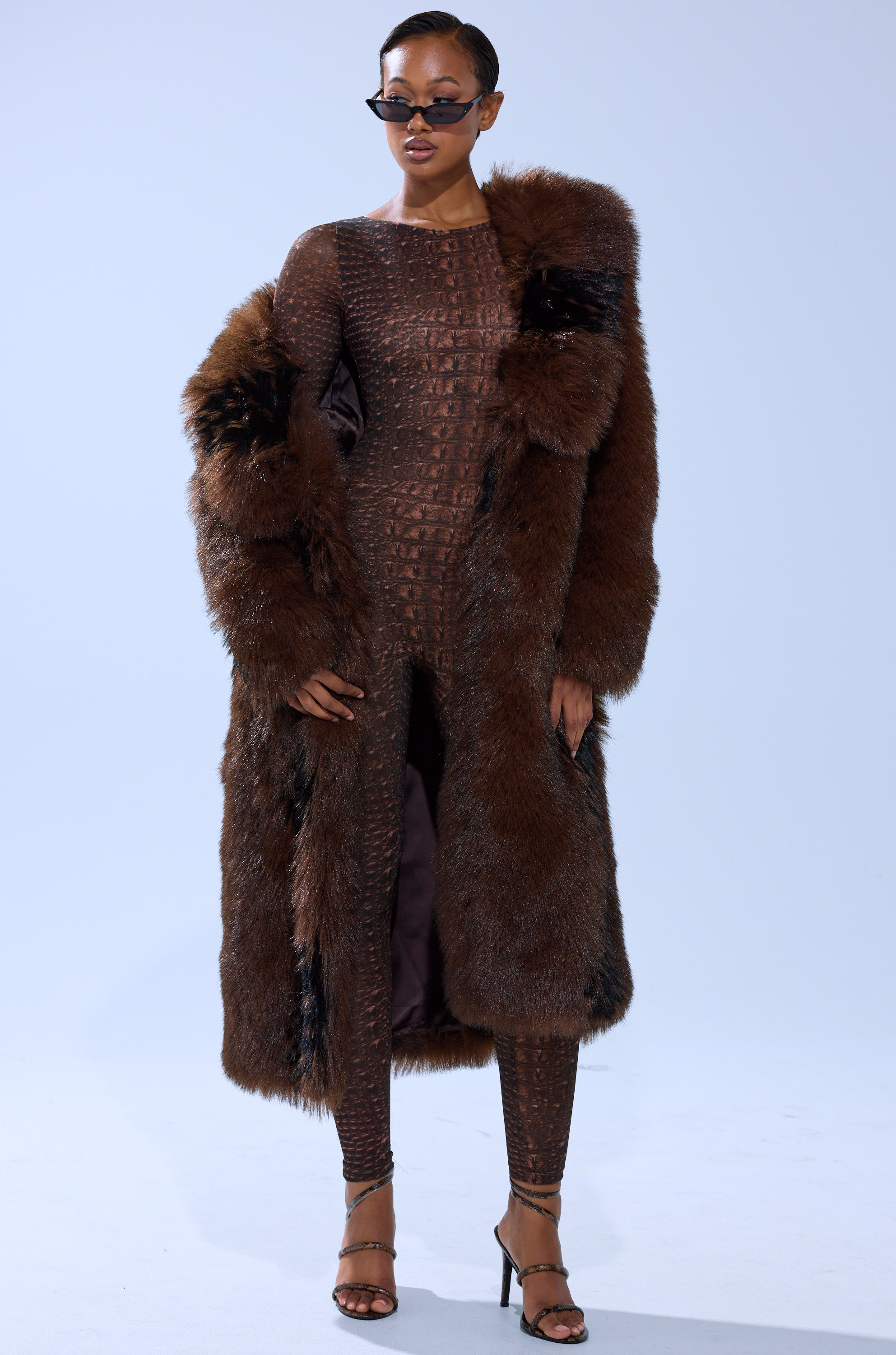 A woman in dark sunglasses models the TELLURIDE FAUX FUR IN BROWN longline jacket over a fitted brown patterned jumpsuit and black strappy heels, set against a plain light background.