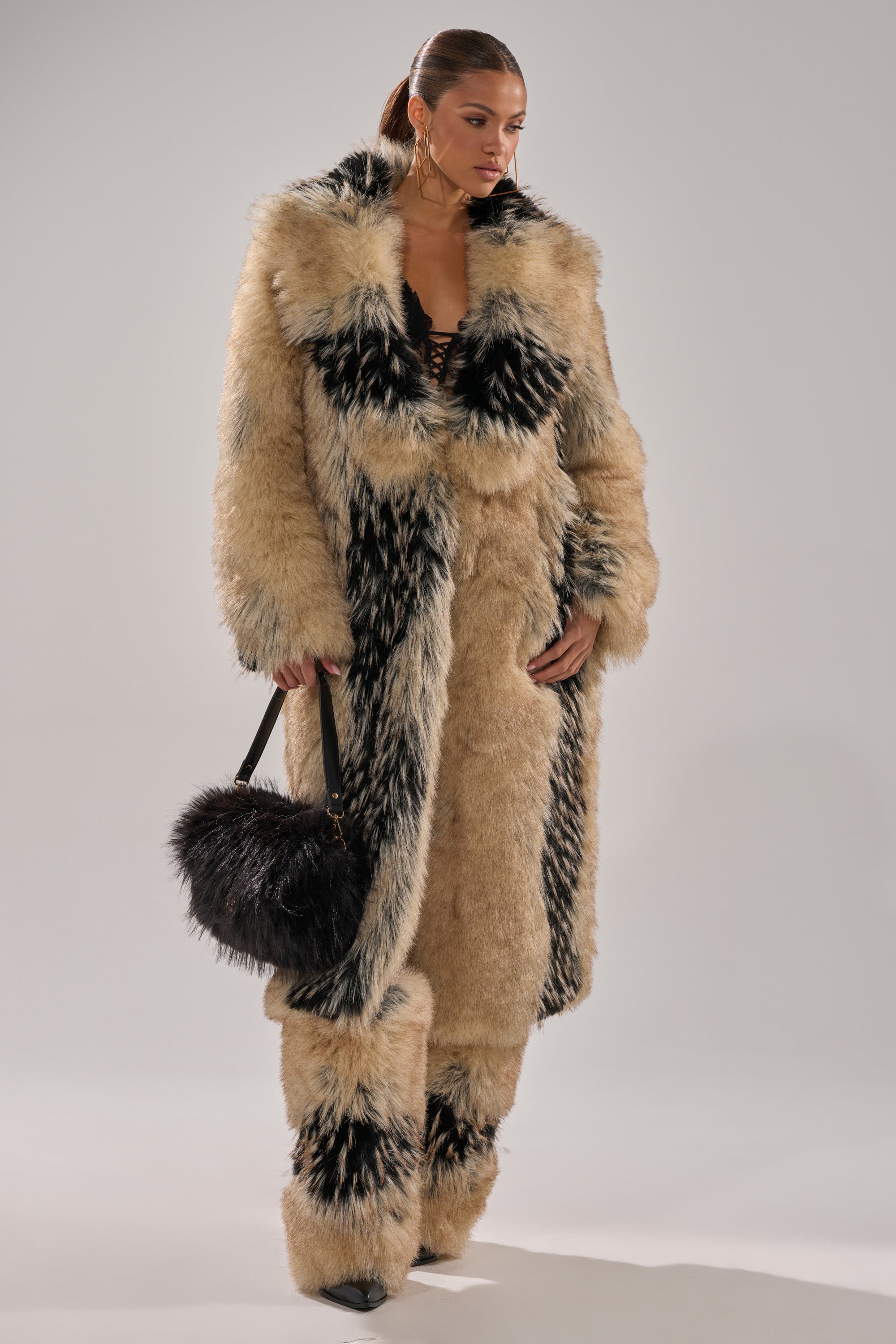 A woman wears the TELLURIDE FAUX FUR IN TAN coat with matching pants and a black handbag, gazing to the side against a plain background—a chic winter essential look.