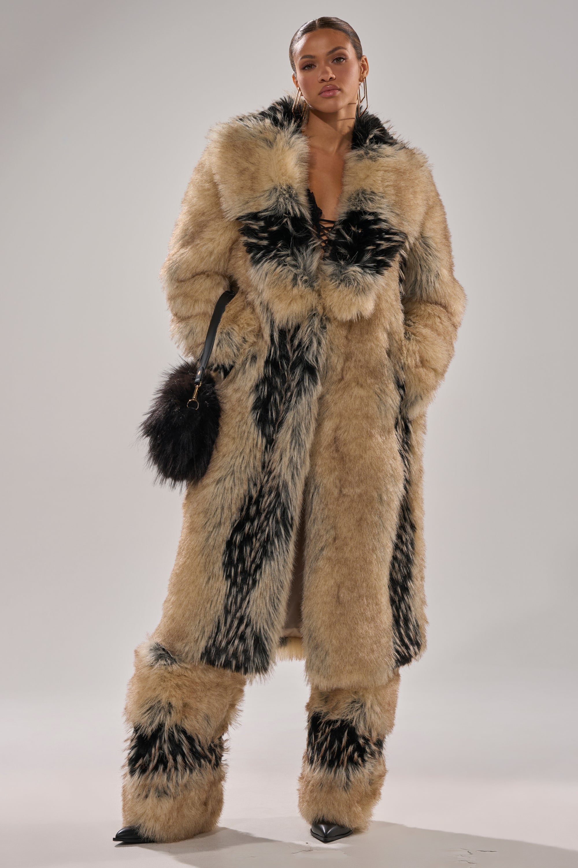 A person stands confidently in the TELLURIDE FAUX FUR IN TAN coat, featuring a longline silhouette, paired with matching wide-leg pants and a black fuzzy handbag, all set against a plain light gray background.