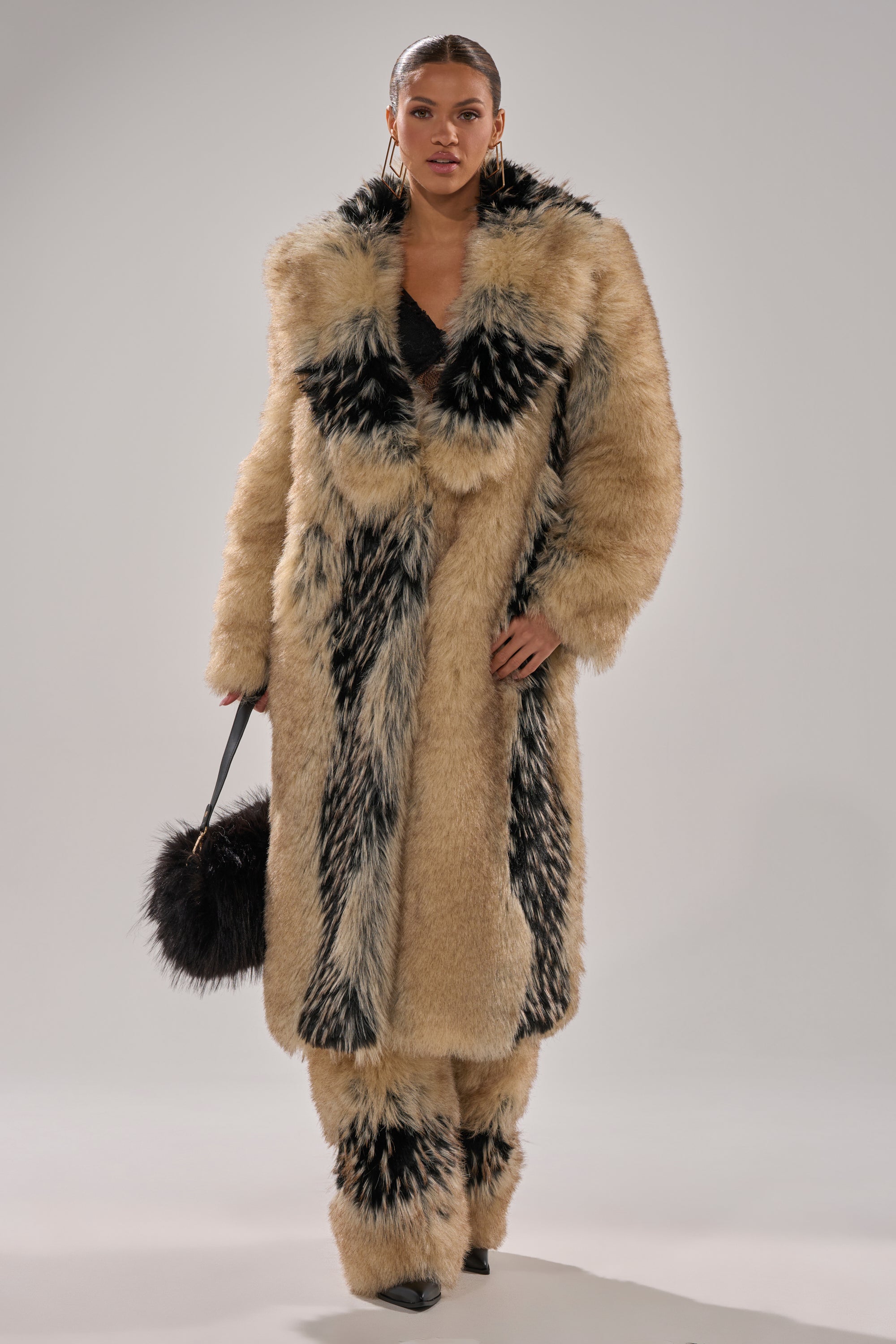 Wearing the TELLURIDE FAUX FUR IN TAN coat with matching pants and black pointed shoes, a person stands against a plain background holding a black furry handbag.
