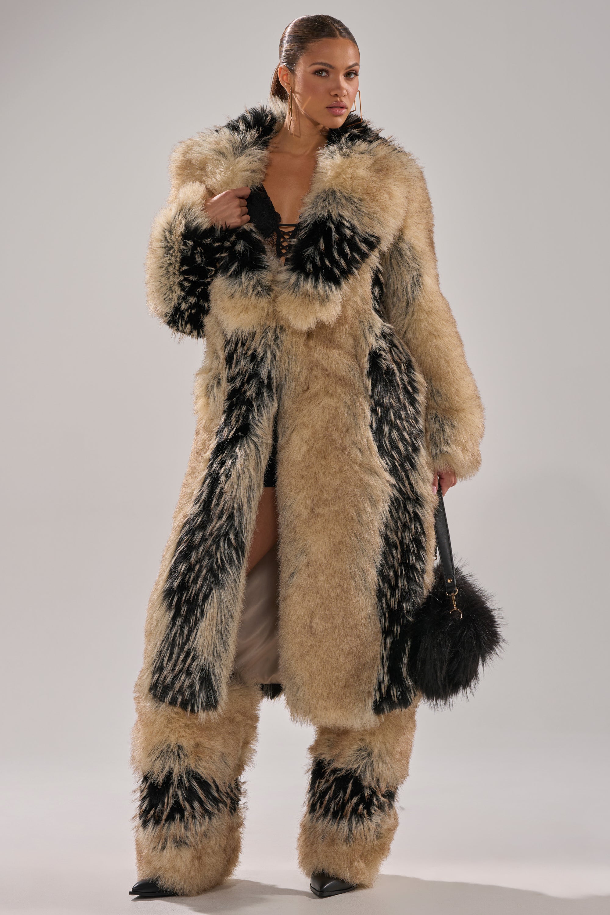 A woman stands confidently in the TELLURIDE FAUX FUR IN TAN, a longline faux fur coat with matching patterned pants and a black furry handbag, her hair pulled back against a neutral background.
