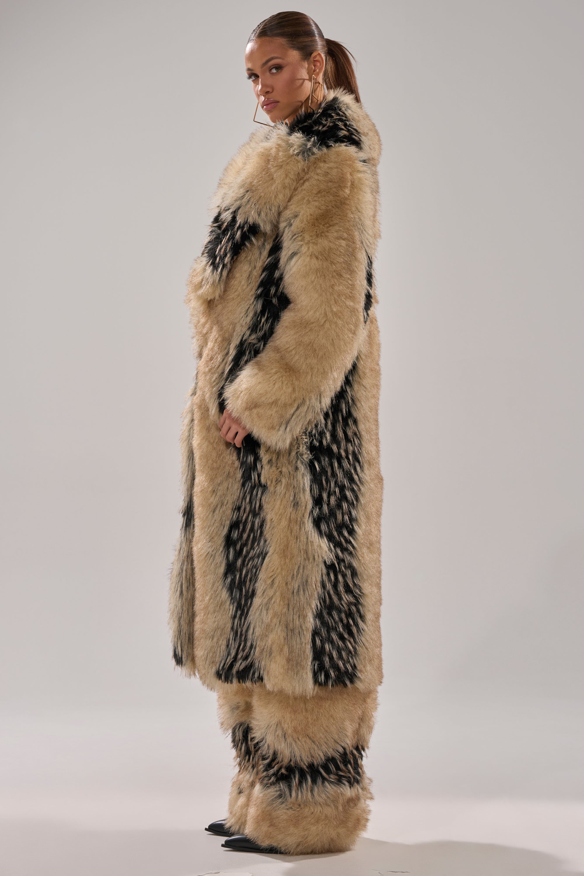 A woman stands sideways in the TELLURIDE FAUX FUR IN TAN coat with matching pants, her hair in a ponytail as she glances over her shoulder against a plain light background.