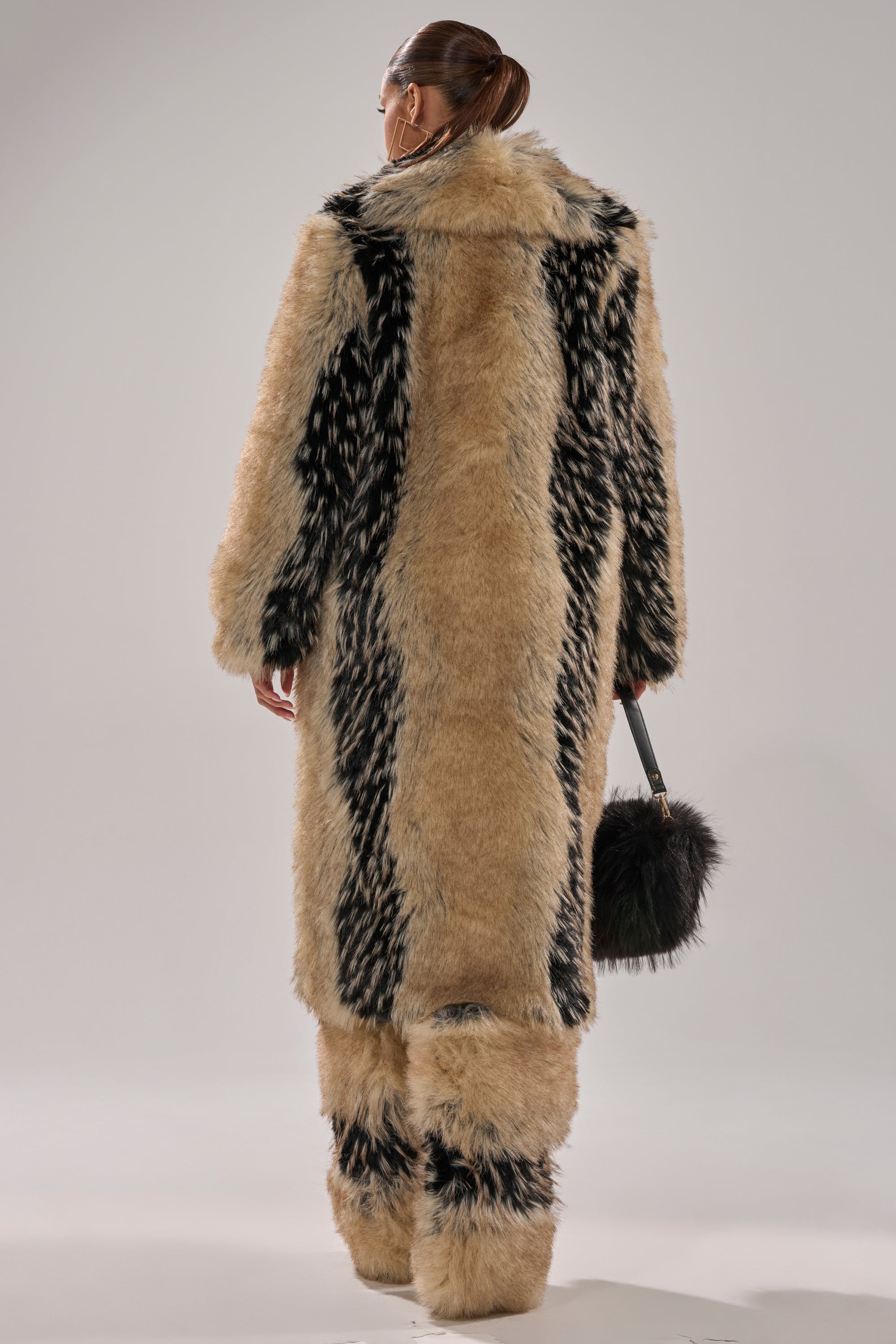 Seen from behind, a person with slicked-back hair wears the TELLURIDE FAUX FUR IN TAN coat and matching pants with a longline silhouette, holding a small round black fur bag against a plain light background.