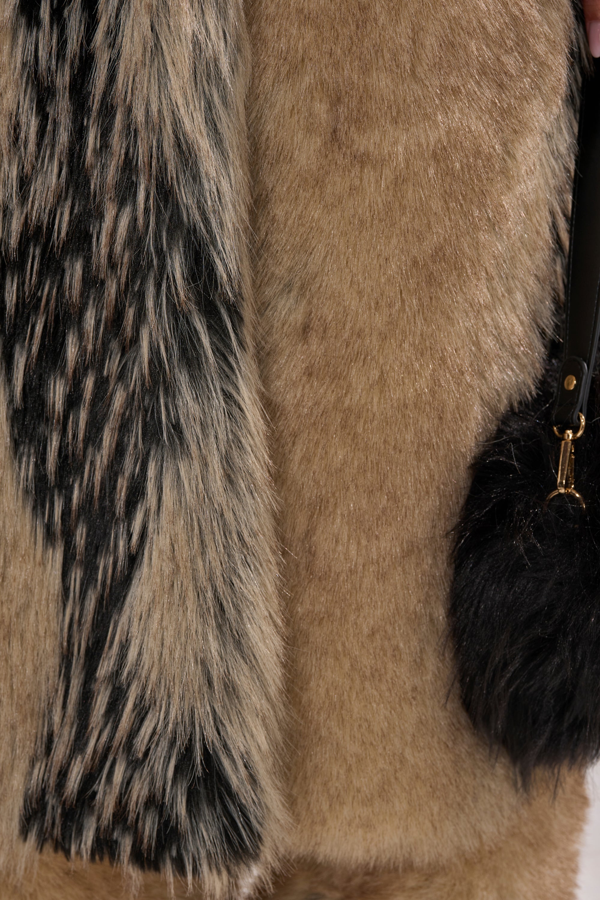 Close-up of a person wearing the TELLURIDE FAUX FUR IN TAN coat, featuring tan with black and white fur accents, holding a black fur accessory with a gold clasp.