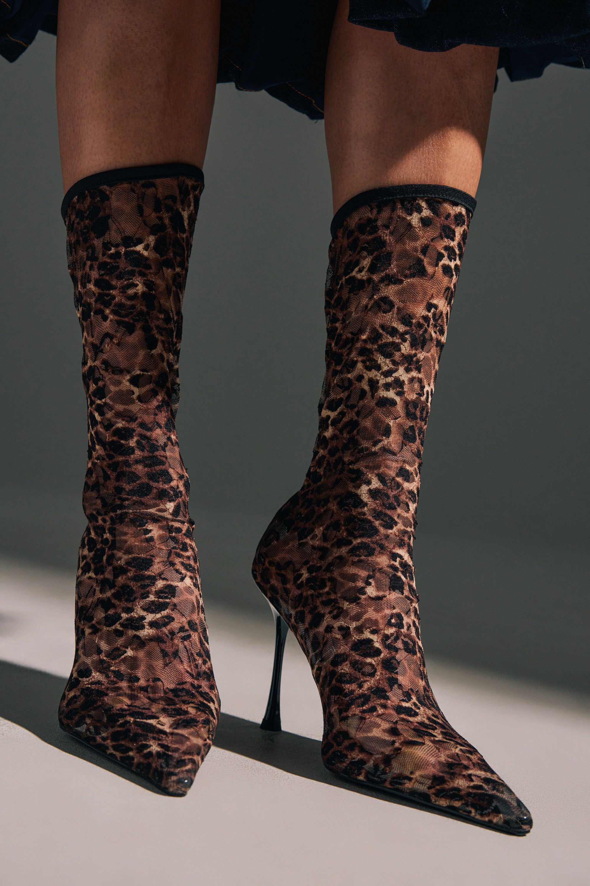 Close-up of legs and feet wearing the AZALEA WANG NICKLAS LEOPARD STILETTO BOOTIE, featuring a pointed toe and mesh upper, standing on a smooth, neutral-colored floor.