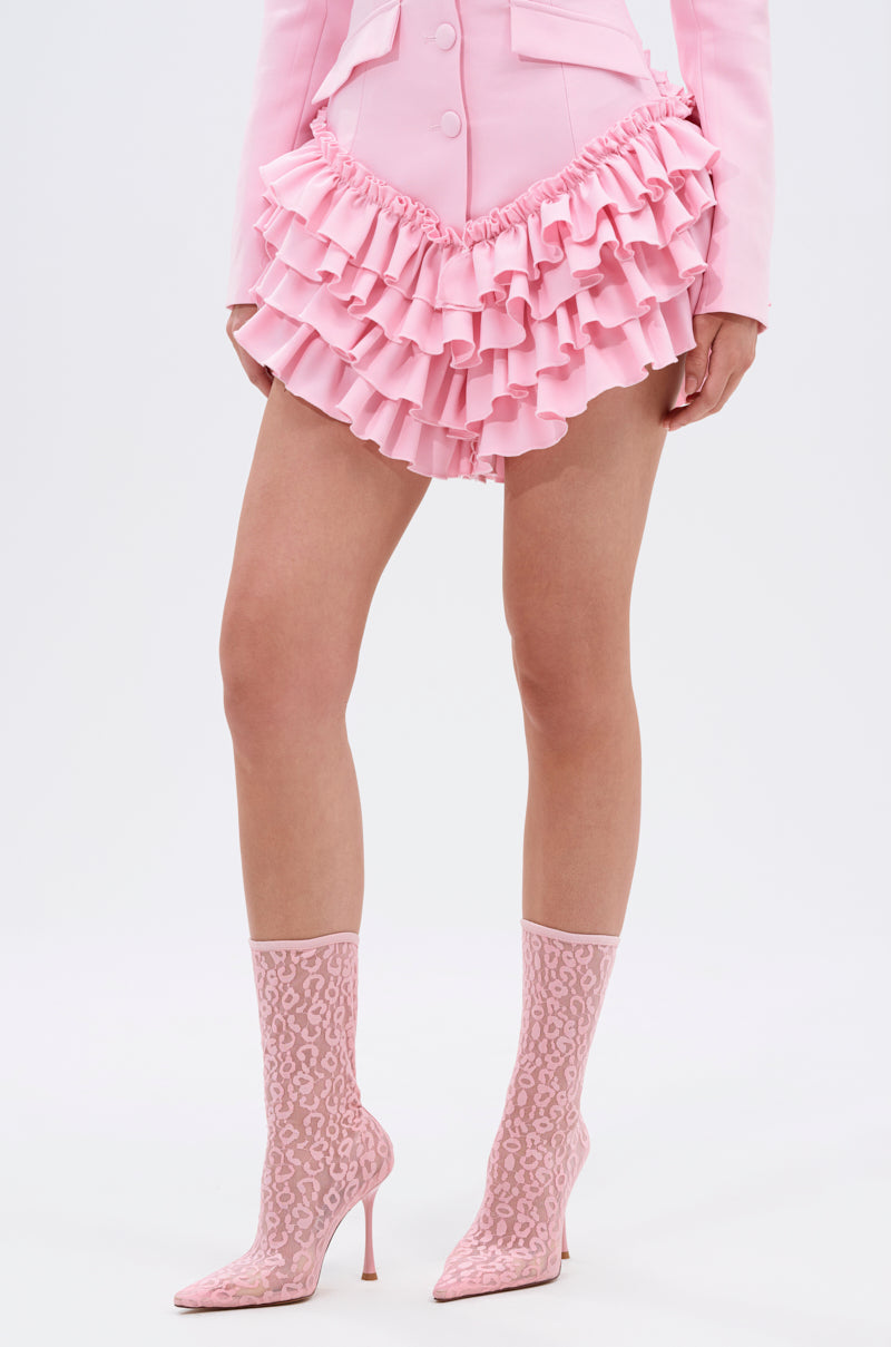 A person in a light pink ruffled mini skirt, matching tailored jacket, and AZALEA WANG NICKLAS PINK LEOPARD STILETTO BOOTIE stands against a plain white background. Their head is not visible in the image.