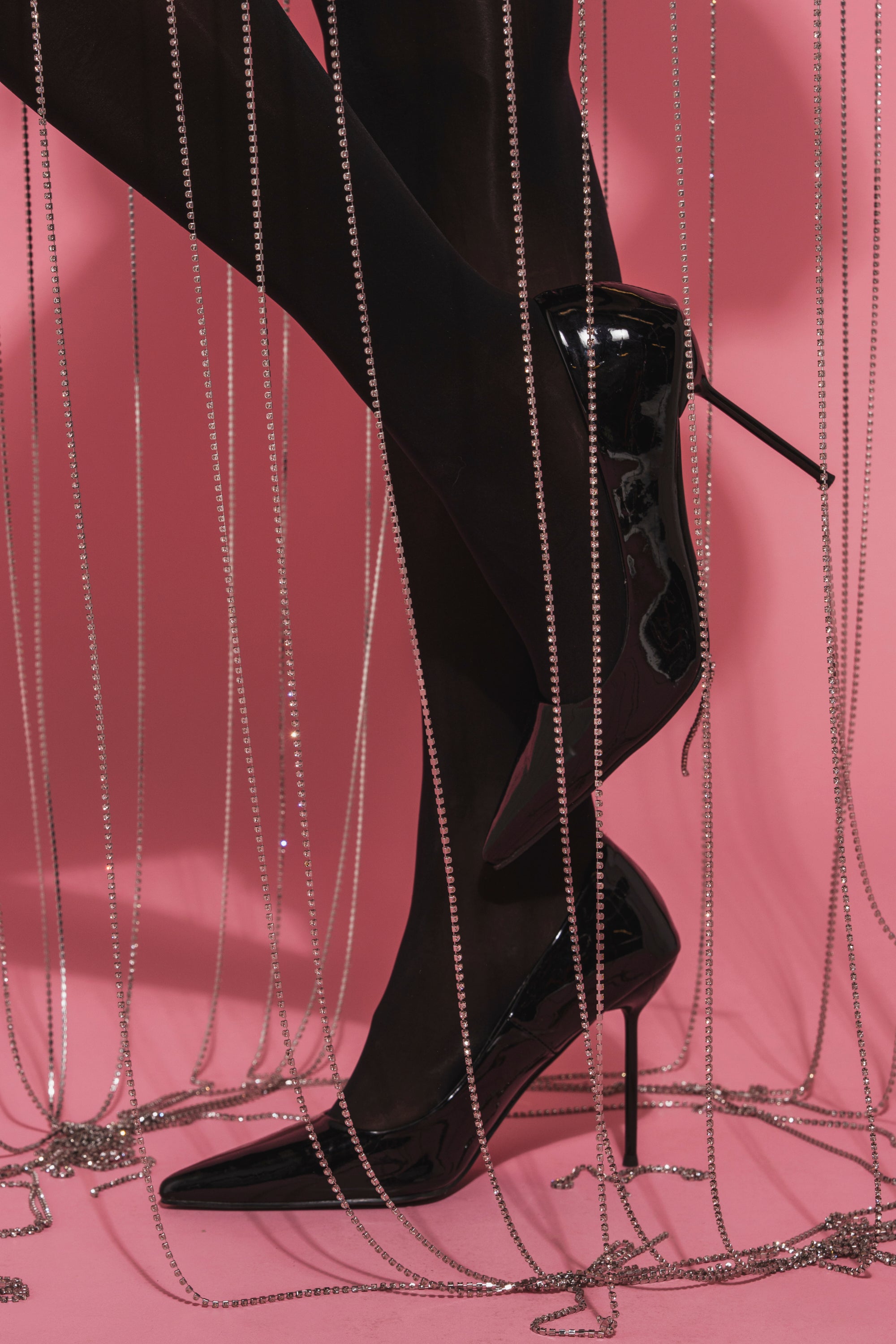 A person wearing the AZALEA WANG NIKOMEDES BLACK PATENT STILETTO PUMP and black tights stands against a pink background, with long, sparkling chains hanging vertically around their legs.