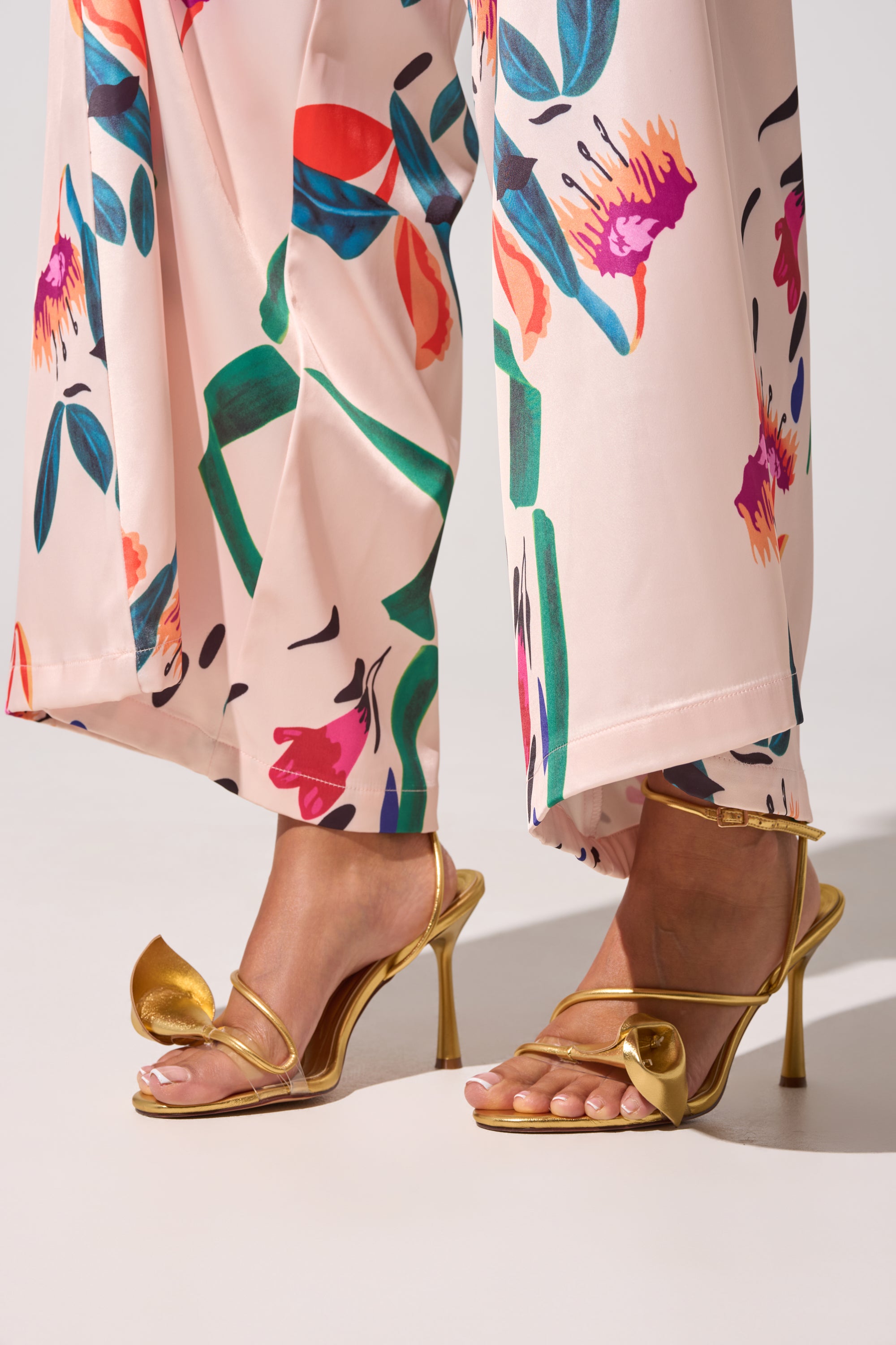 A person in wide-legged, light pink floral pants showcases the AZALEA WANG NILO GOLD METALLIC FLORAL DECALE STRAP STILETTO SANDAL, styled perfectly for standout special occasion footwear. 0
