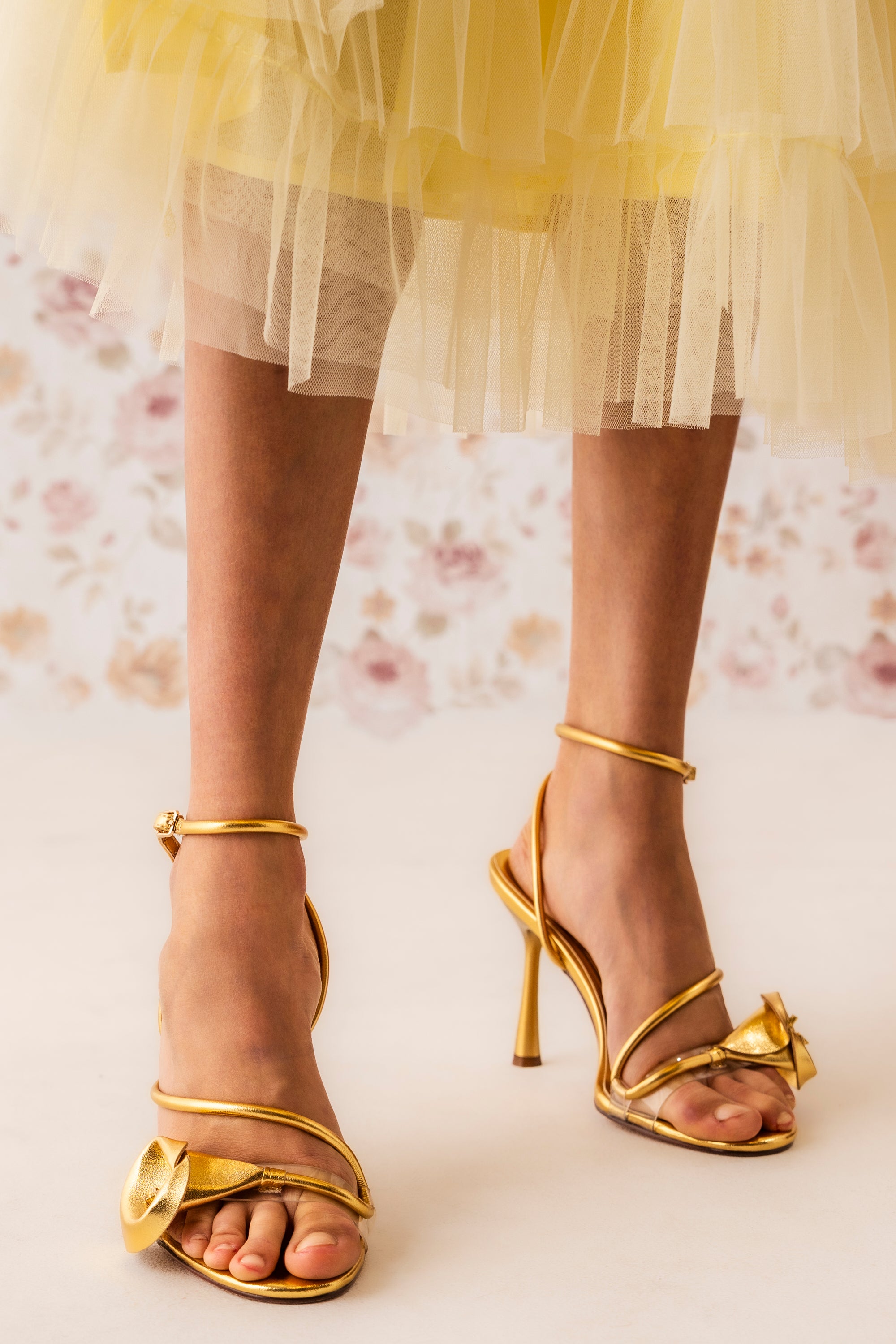 Wearing the AZALEA WANG NILO GOLD METALLIC FLORAL DECALE STRAP STILETTO SANDAL, a person stands on a light surface in a yellow tulle skirt, set against a soft floral background. 1