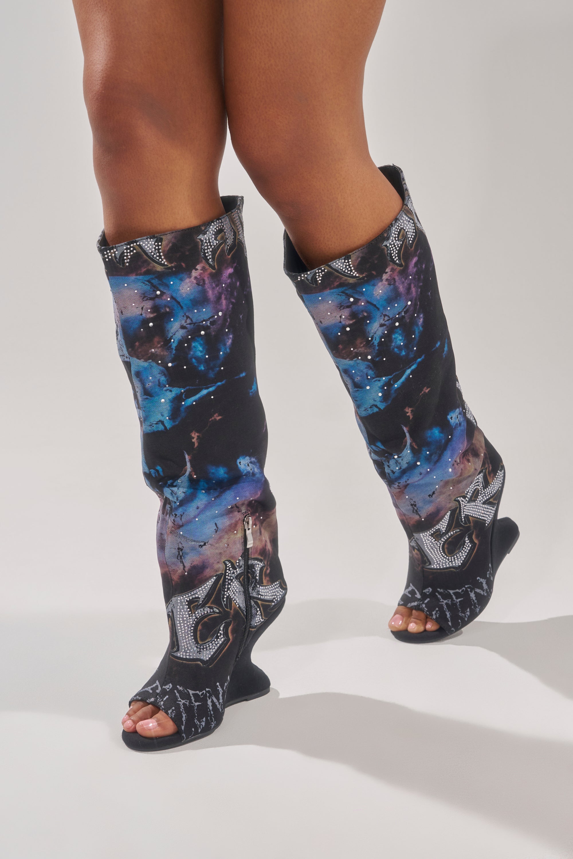A person wearing the AKIRA GOLD NINE LIVES GRAPHIC OPEN TOE WEDGE BOOT—knee-high boots with a galaxy print and silver embellished letters on white.