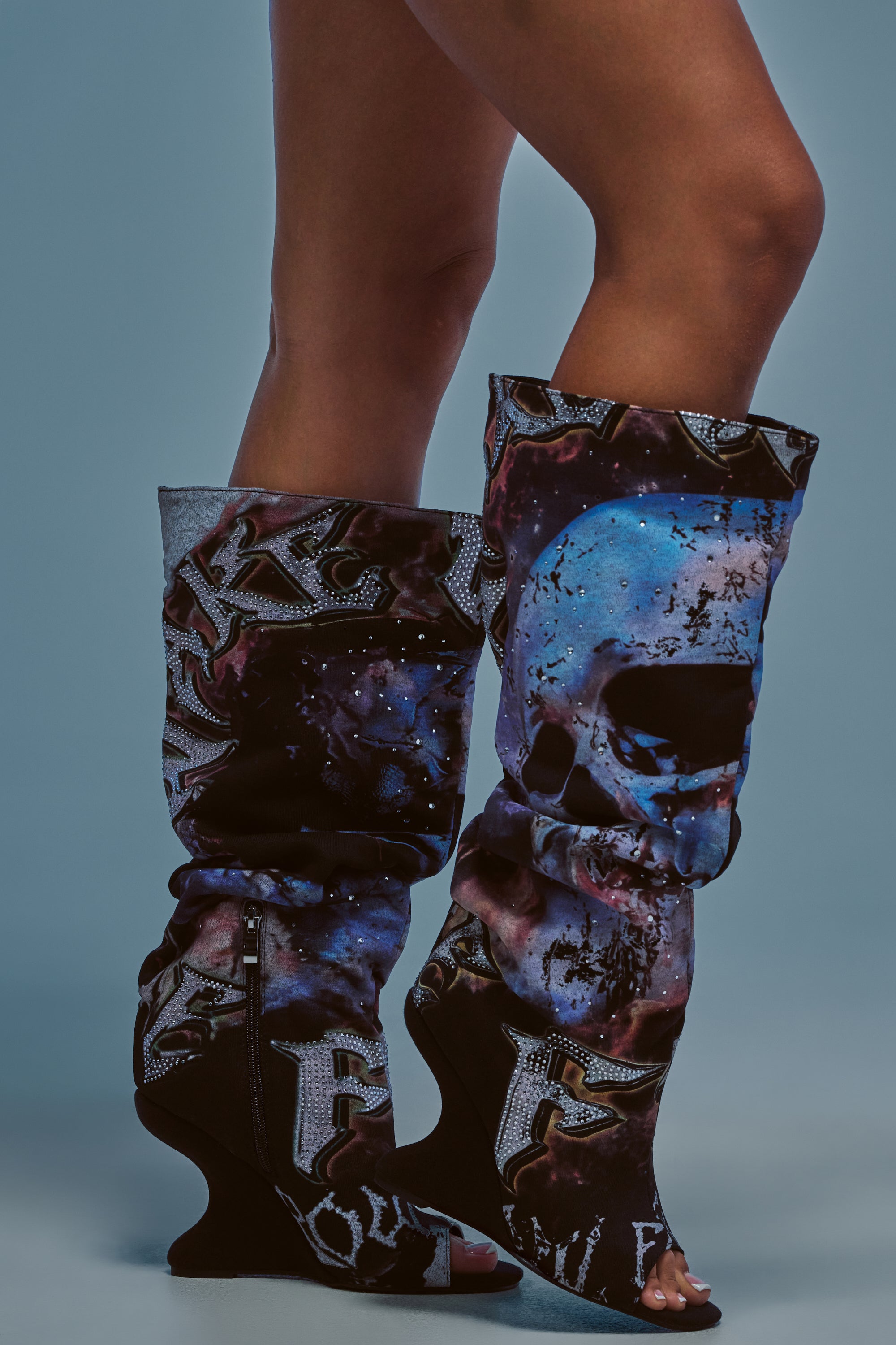 A person wearing the AKIRA GOLD NINE LIVES GRAPHIC OPEN TOE WEDGE BOOT stands against a blue background. The knee-high boots showcase a cosmic, skull-inspired design with subtle Y2K details and an open toe, with only the legs visible.