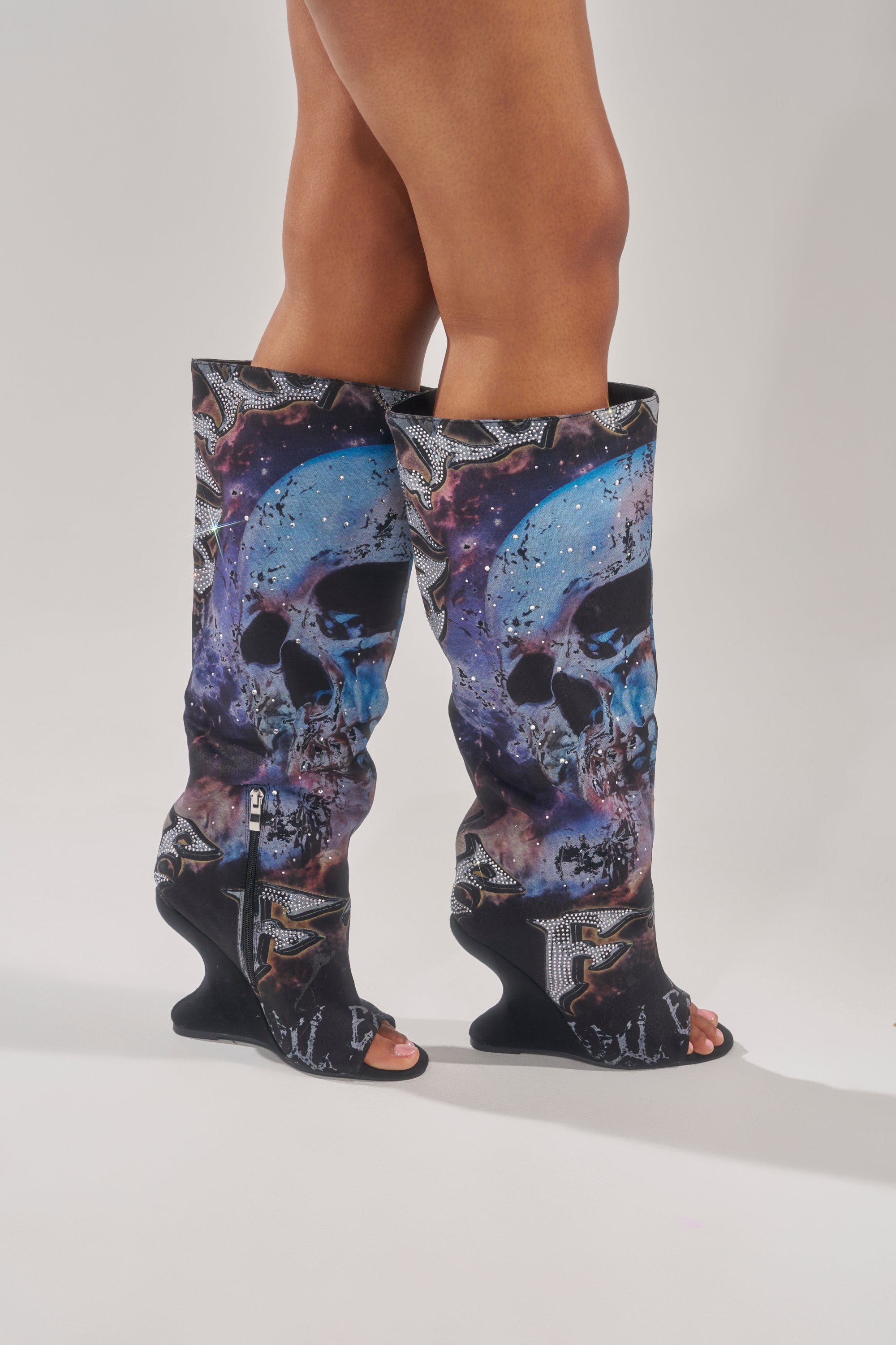 A person models the AKIRA GOLD NINE LIVES GRAPHIC OPEN TOE WEDGE BOOT—knee-high boots with a bold blue and purple skull print, side zippers, sculpted heels, and open toes—against a plain background.