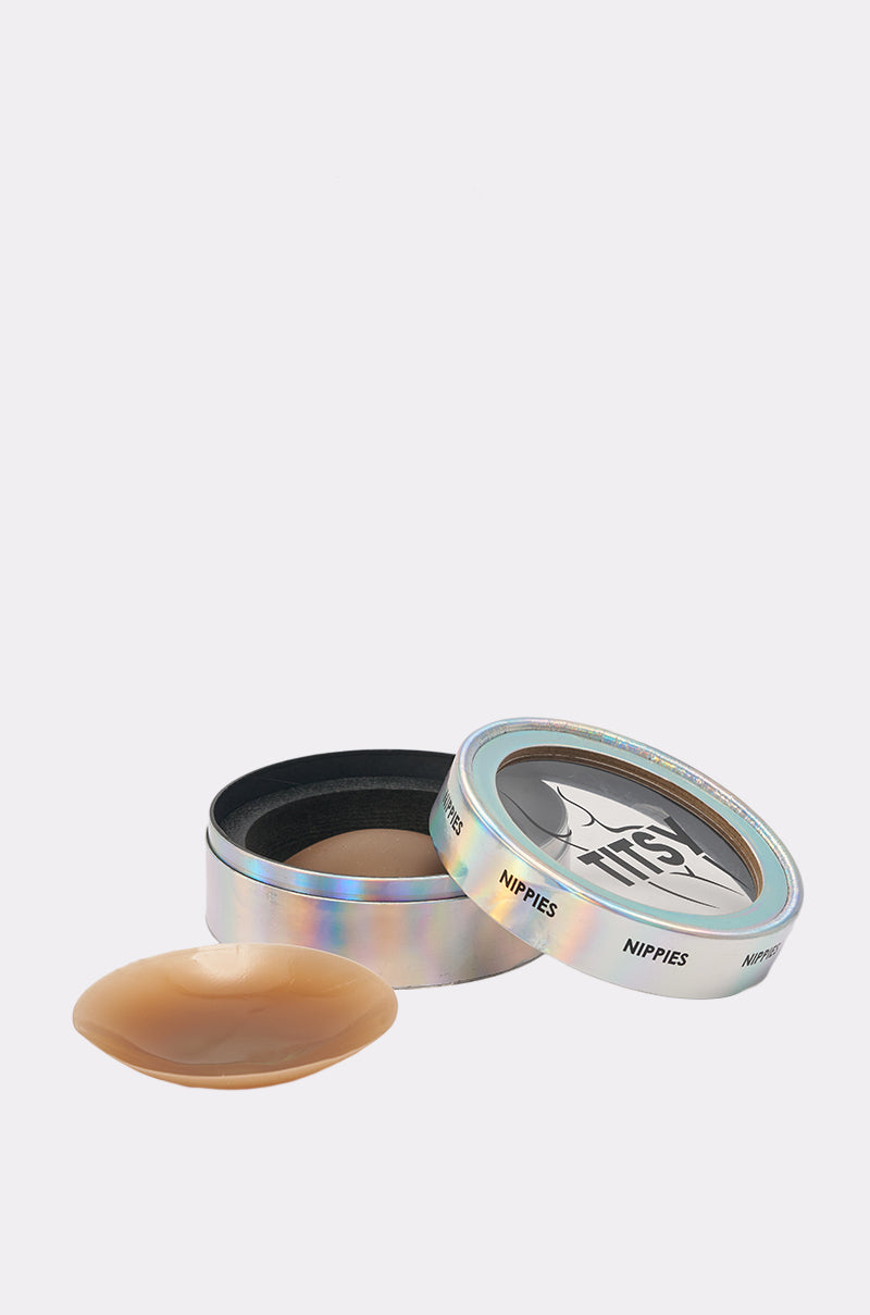 A pair of nude TITSY ADHESIVE SILICONE NIPPIE PETALS, with one in a round iridescent tin labeled “Nippies” and the other displayed in front on a white background. The tin highlights discreet adhesive nipple covers. 0