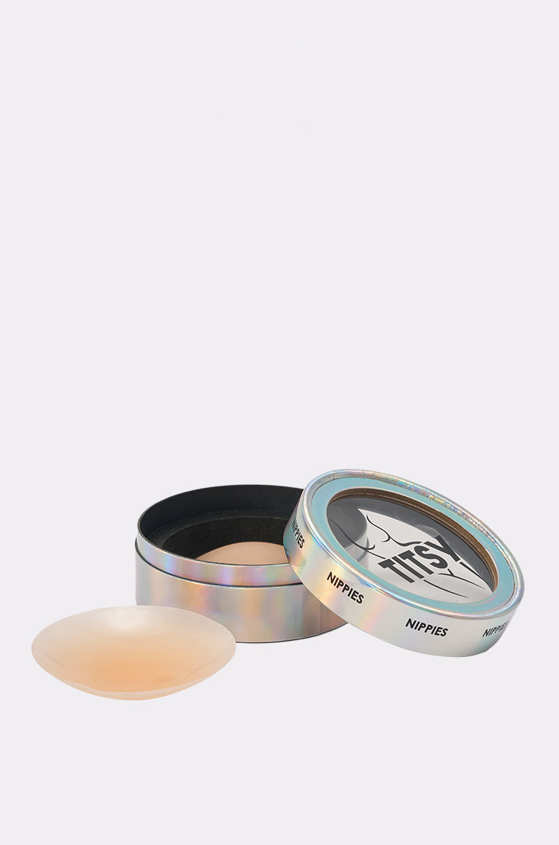 An open, round iridescent tin labeled "NIPPIES" with a clear lid reveals TITSY ADHESIVE SILICONE NIPPIE PETALS, including one seamless nude silicone cover displayed outside the container. 0