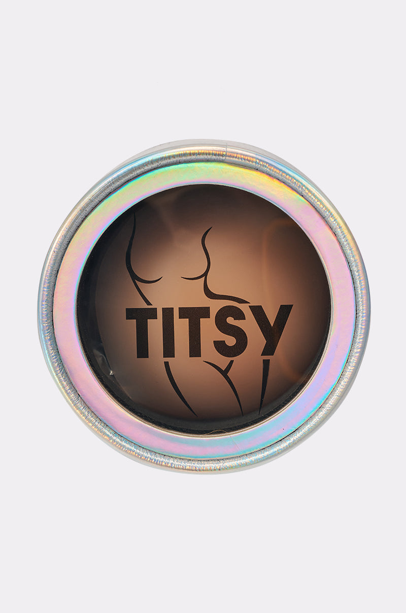 A round, holographic tin with a clear lid displays "TITSY" and a minimalist torso design on brown—a stylish case for storing TITSY ADHESIVE SILICONE NIPPIE PETAL nude silicone nipple covers. 1
