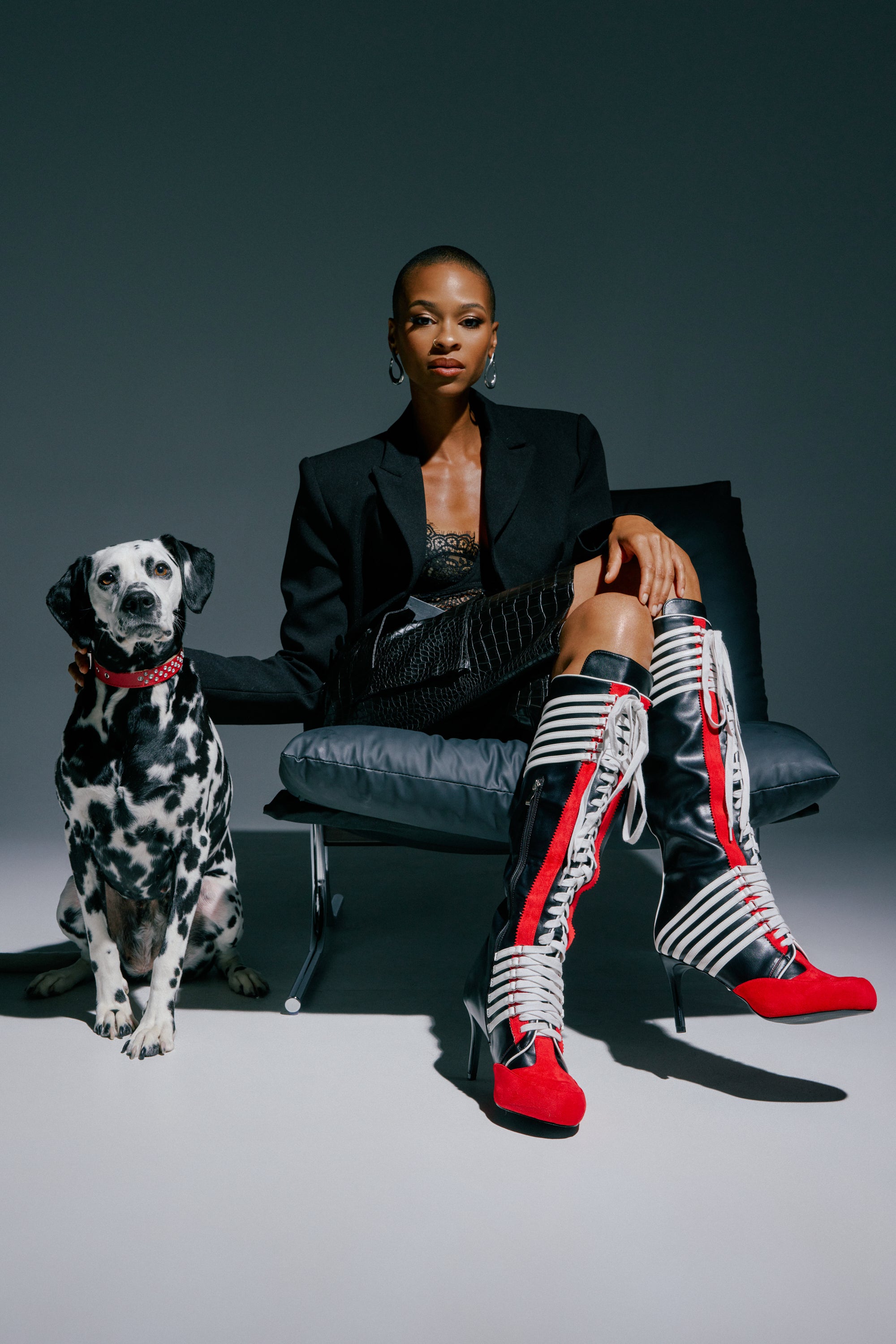 A woman dressed in black sits on a chair next to a Dalmatian wearing a red collar, both posed against a dark, minimalist background; she completes her look with the AZALEA WANG NITRO RED MULTI SPORTY STILETTO BOOT featuring varsity-inspired color blocking.