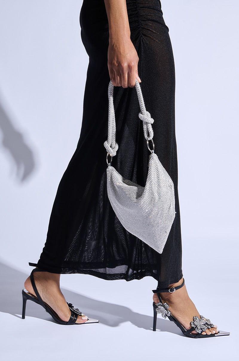 A person wearing a black dress and AZALEA WANG NJEMA FLOWER EMBELLISHED STILETTO SANDALS holds a small, silver mesh handbag with large knots against a plain white background.