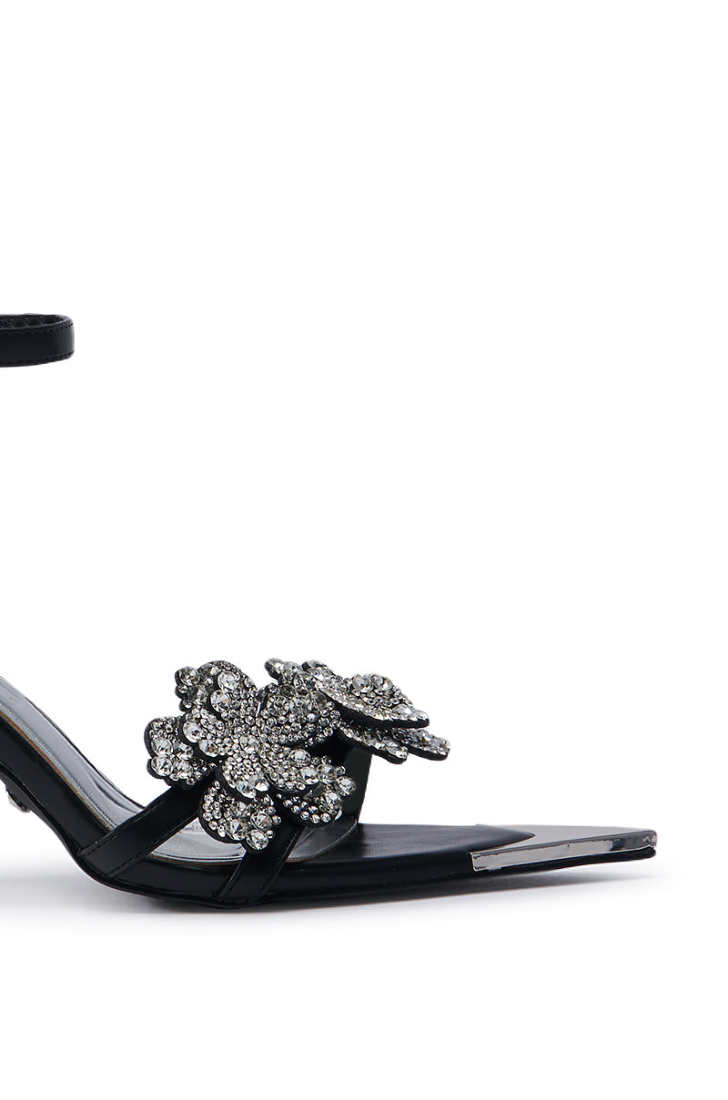 The AZALEA WANG NJEMA FLOWER EMBELLISHED STILETTO SANDAL is a black high-heeled shoe featuring an ankle strap and pointed toe, adorned with flower-embellished stiletto accents and rhinestone floral appliques. It is showcased against a plain white background.