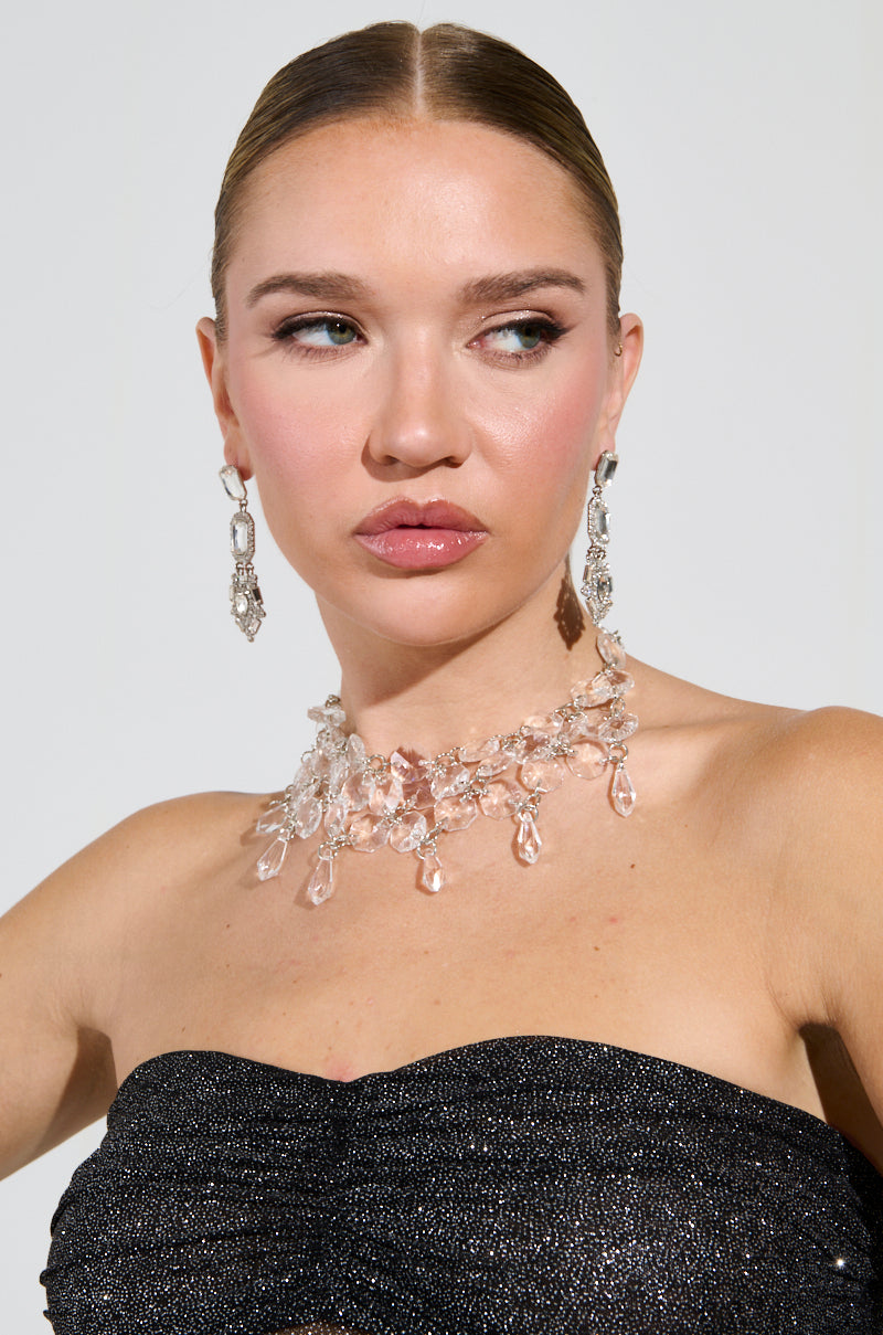 A woman with slicked-back hair, glittering drop earrings, and a FANTASY DRIP CHOKER stands elegantly. In a shimmering black strapless top, she gazes sideways against a plain backdrop, exuding elegance and allure.