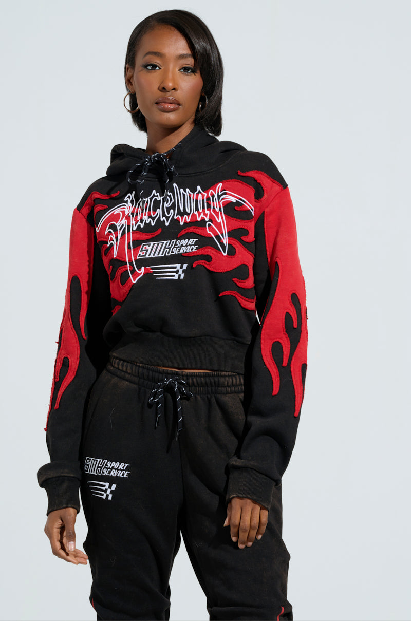 A person stands confidently in an ON FIRE SWEATSHIRT, a black and red hoodie adorned with racing-inspired text and embroidered flame designs. They are paired with matching black pants that feature bold white text on one leg, set against a plain, light-colored background.