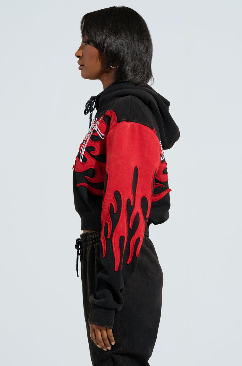 A person in profile showcases the ON FIRE SWEATSHIRT, highlighting its embroidered flame designs on a black and red hoodie with matching pants. The detailed red patterns ignite the sleeves and chest, as their shoulder-length hair cascades against a plain white background.
