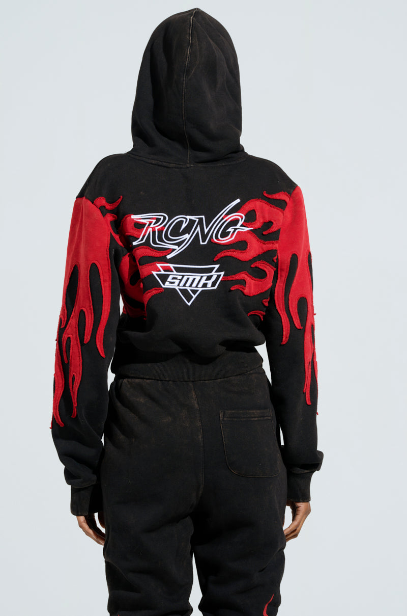 A person wears the ON FIRE SWEATSHIRT, a black hooded masterpiece featuring racing-themed text on the back that reads "RCNG" and "SMTH." The sleeves showcase bold embroidered flame designs, perfectly complemented by matching black pants.