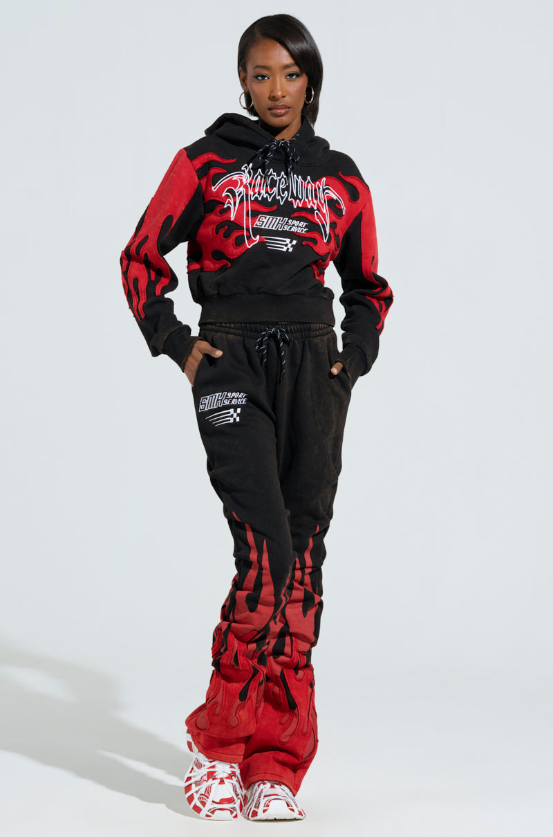 The person sports an eye-catching red and black tracksuit featuring racing-inspired text. The outfit includes a hoodie and sweatpants adorned with embroidered flame designs reminiscent of the ON FIRE SWEATSHIRT, all set against a plain background.
