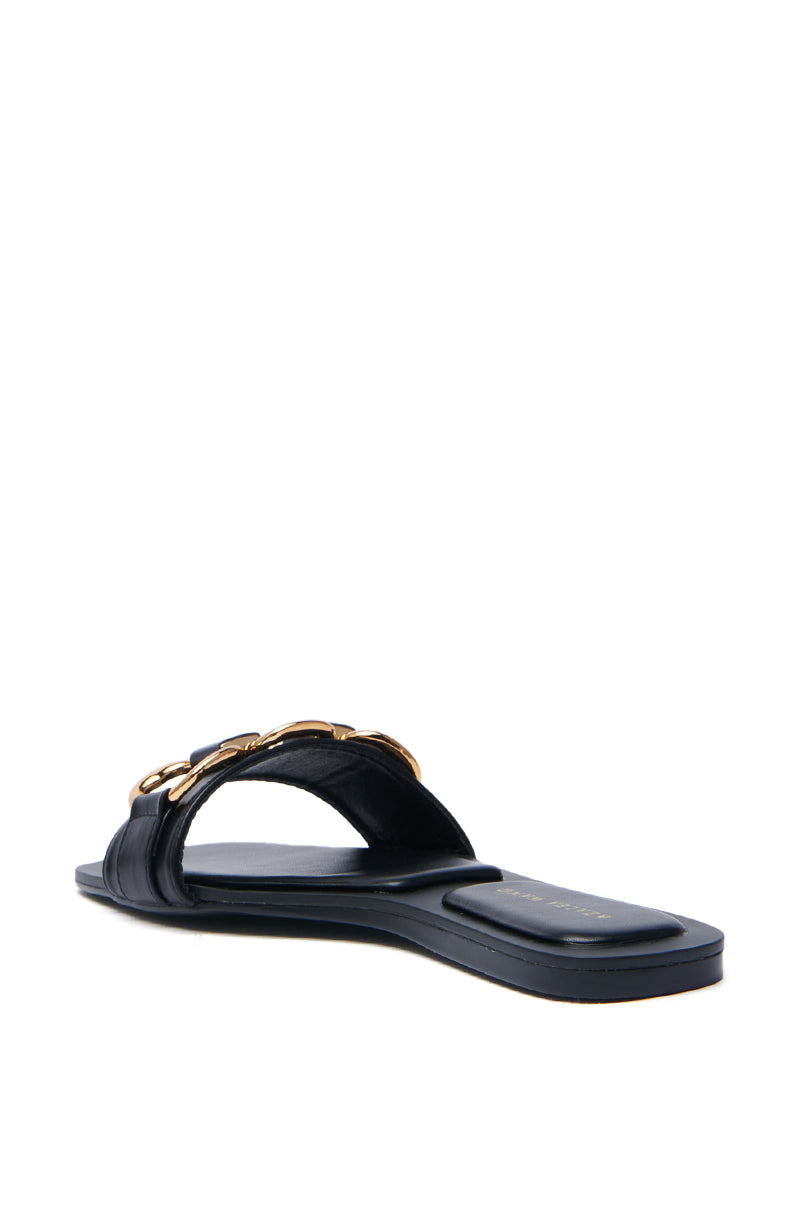 The AKIRA NOELINE black slip-on sandal features a flat sole, wide faux leather strap, and elegant gold chain detail, partially revealing the footbed.