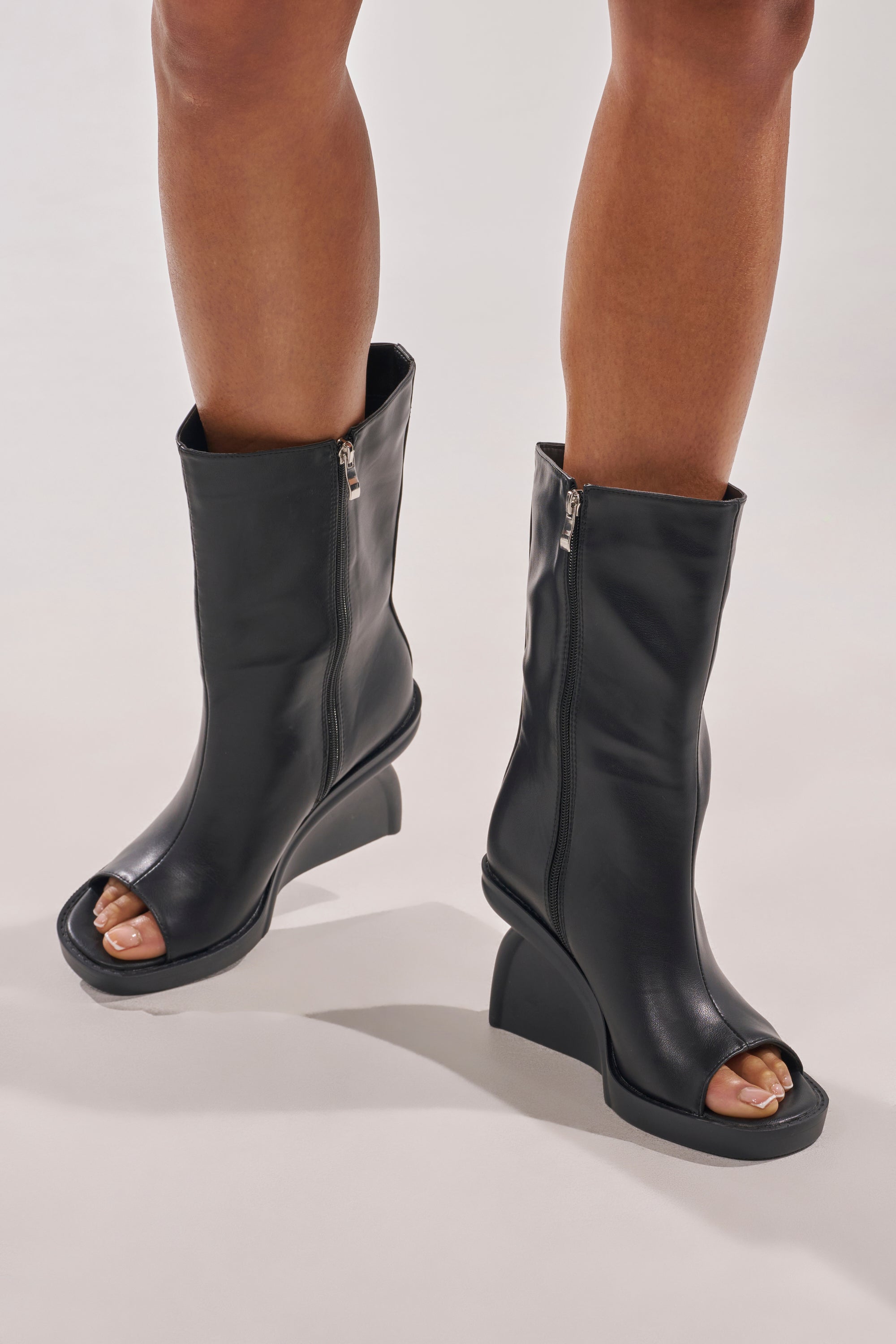 A person stands on a light surface wearing the AZALEA WANG NOLIA BLACK NOVELTY WEDGE BOOTIE, featuring black faux leather, a chunky wedge heel, and side zippers. 1