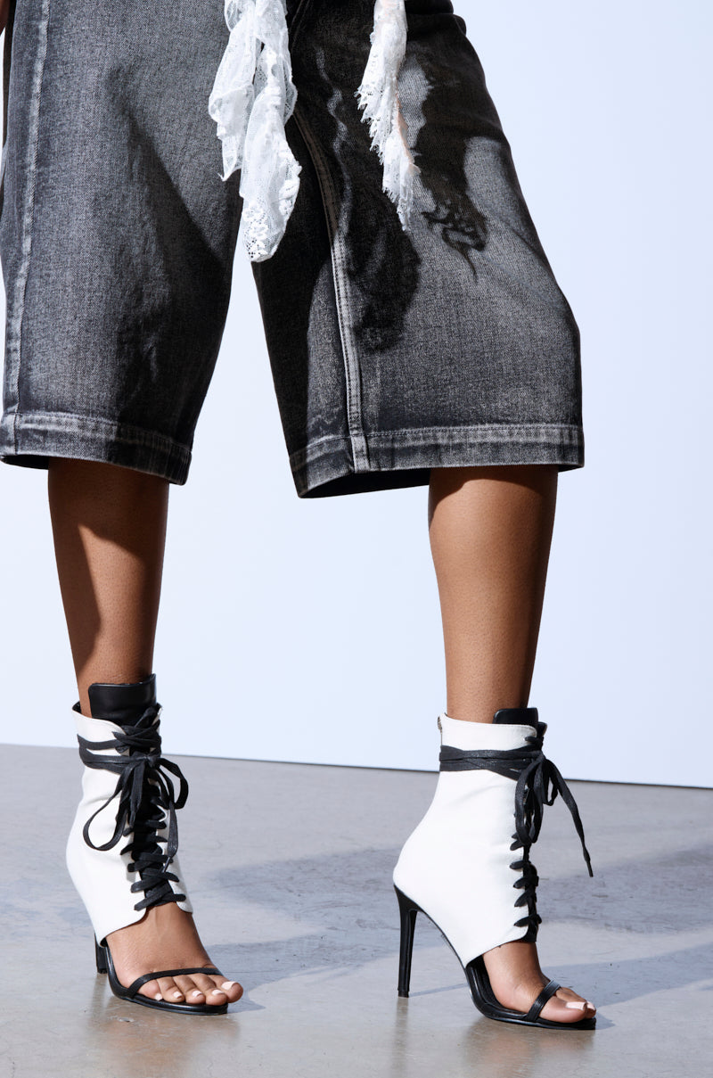 Close-up of a person wearing AZALEA WANG NOLYN HEELED SANDAL, featuring black and white faux leather with open toes and stiletto heels. They pair the sandals with dark denim shorts and a flowing white lace garment against a light background.