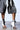 Close-up of a person wearing AZALEA WANG NOLYN HEELED SANDAL, featuring black and white faux leather with open toes and stiletto heels. They pair the sandals with dark denim shorts and a flowing white lace garment against a light background.