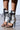 A person showcases the AZALEA WANG NOLYN HEELED SANDAL, featuring a sleek stiletto heel, white design with black leather laces and trim, and an open-toed, lace-up mid-calf shaft against a neutral light-colored floor.
