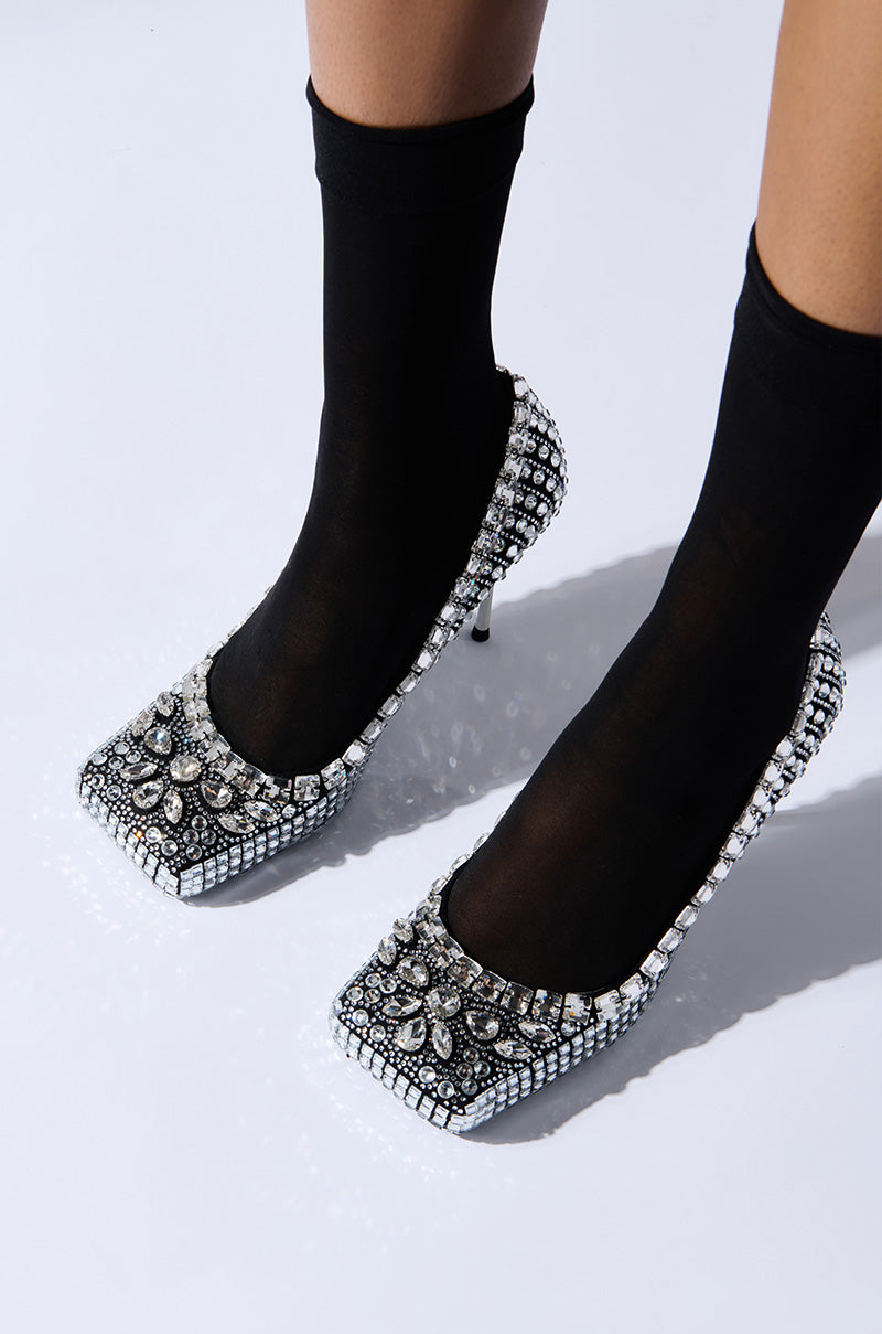 A person wearing the AZALEA WANG NOOMI BLACK PUMP with black socks. The shoes display a square toe, intricate floral designs, and are adorned with numerous silver rhinestones, exuding a luxurious and glamorous charm.