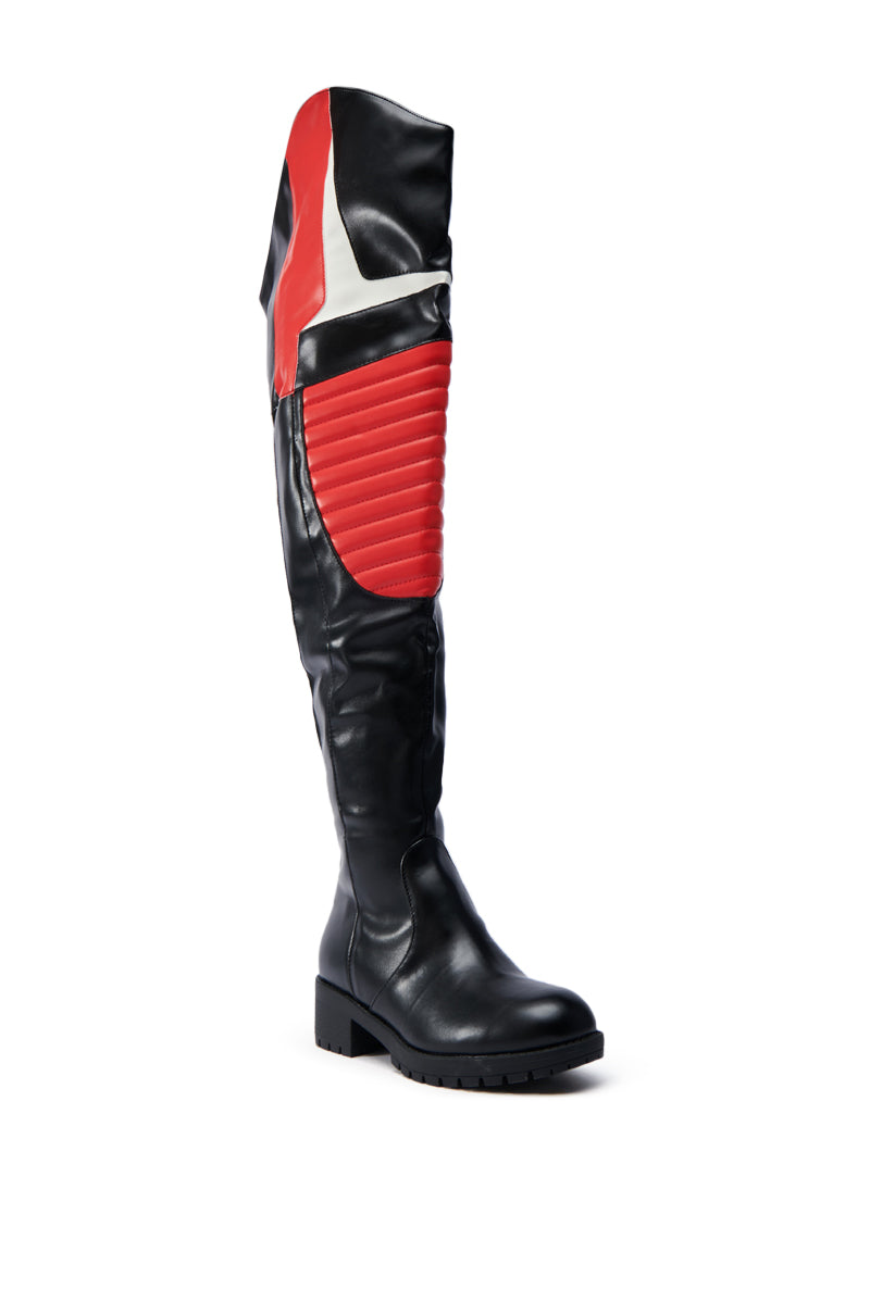 The NORRIS RED AND BLACK FLAT BOOT is a thigh-high moto boot with a textured red panel, smooth and ribbed faux leather, and red and white accents. It features a flat rubber lug sole and extends over the knee.