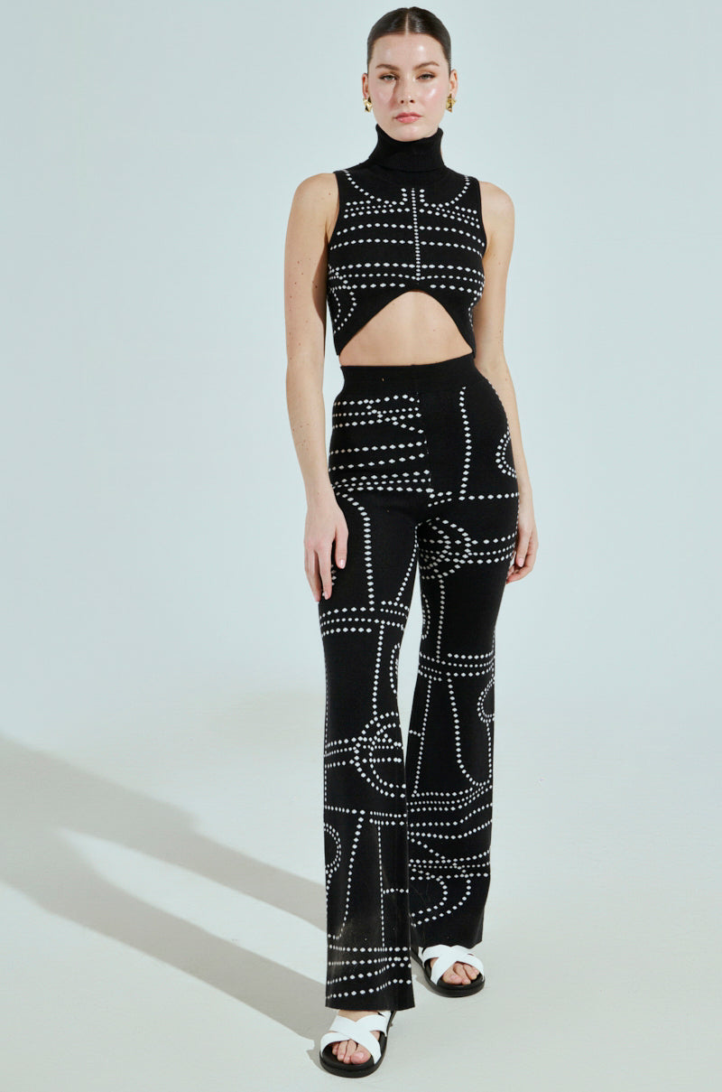 A person is in a black sleeveless turtleneck with a front cutout, paired with high-waisted "DOTTED MINI FLARE PANT," all in white dotted knit. They complete the look with white open-toe sandals, set against a light gray background.