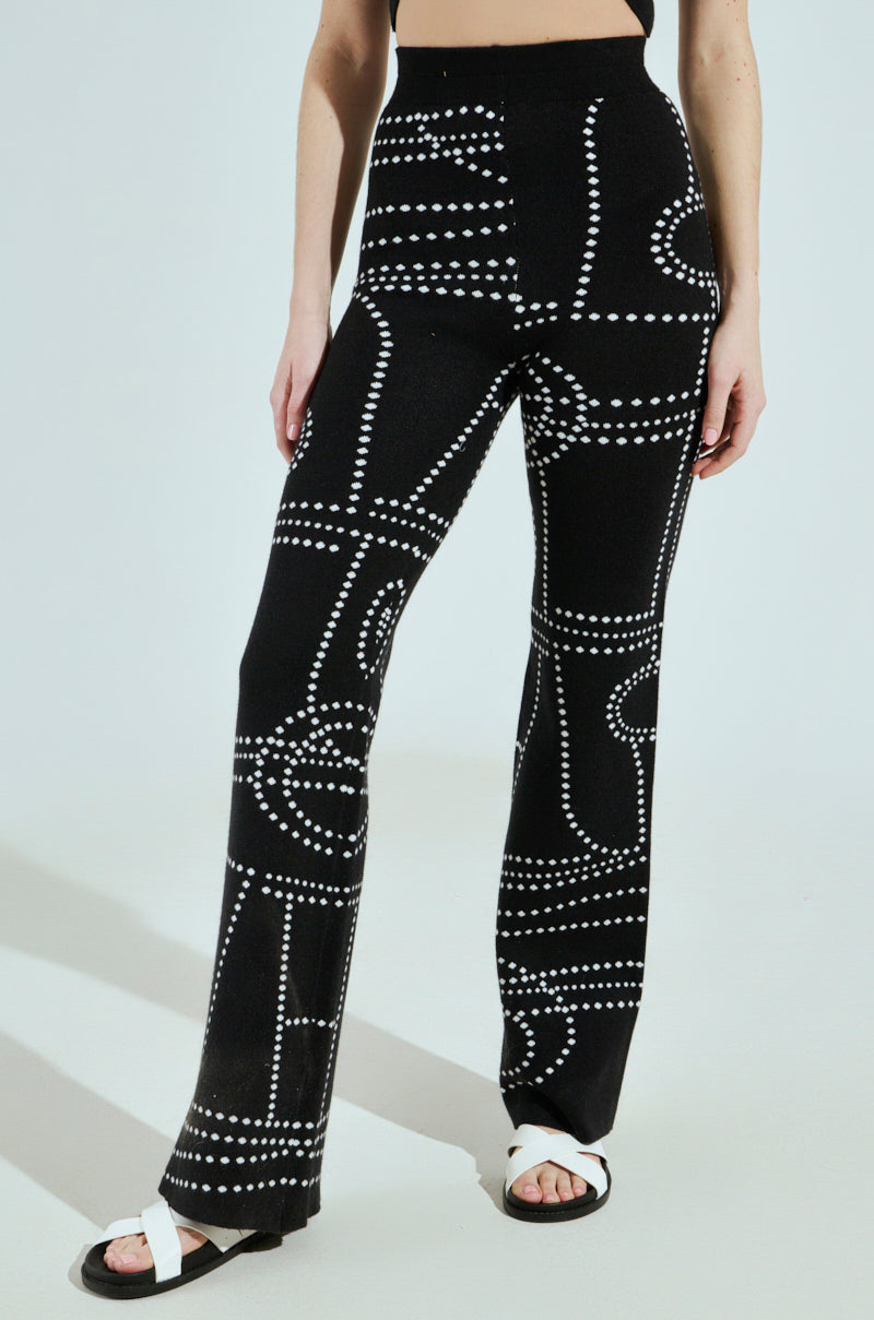 The individual is wearing Dotted Mini Flare Pants in a white abstract pattern with a fitted knit design and flared legs, paired with a black top. The outfit is completed with white sandals featuring black straps on a plain, light-colored backdrop.