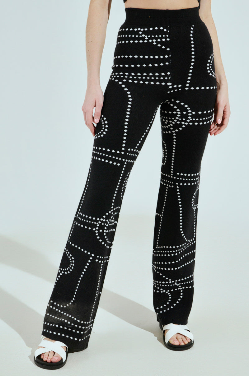 A person stands in high-waisted black pants with a white dotted geometric pattern, similar to the DOTTED MINI FLARE PANT. Their hands are by their side, they wear white sandals, and they're against a plain light background.
