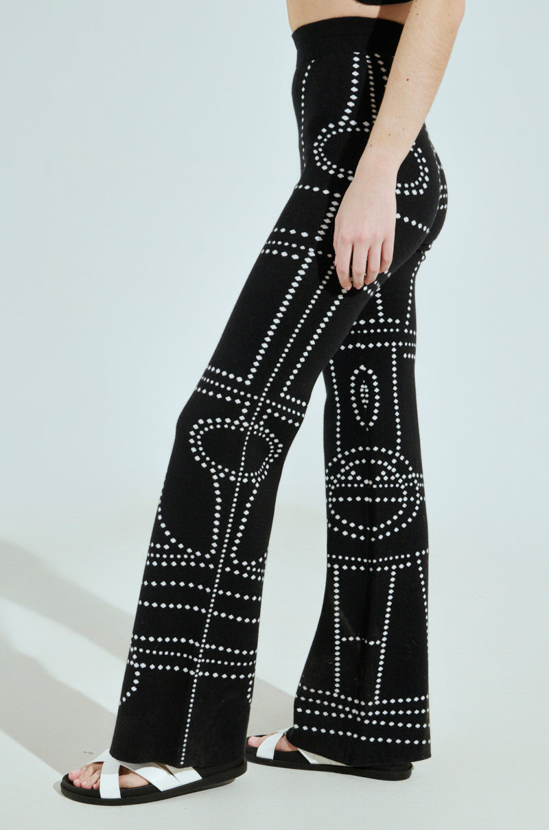 A person is shown wearing black DOTTED MINI FLARE PANT with a geometric pattern of white dots. These knit fabric pants are paired with white sandals, focusing on the lower body against a plain background.