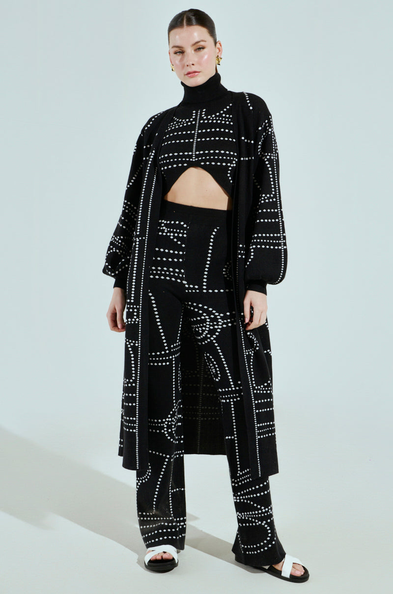 A person exudes an artsy vibe against a light gray background, wearing a black outfit with a dotted pattern. The look includes the DOTTED OVERSIZED CARDIGAN, pants, and a high-neck top with a cut-out section. White sandals complete the ensemble.