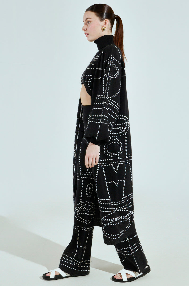 A person exudes an artsy vibe in a DOTTED OVERSIZED CARDIGAN paired with matching pants. Black sandals with white bands complement the look, and their hair is tied back in a ponytail against a pale blue background.