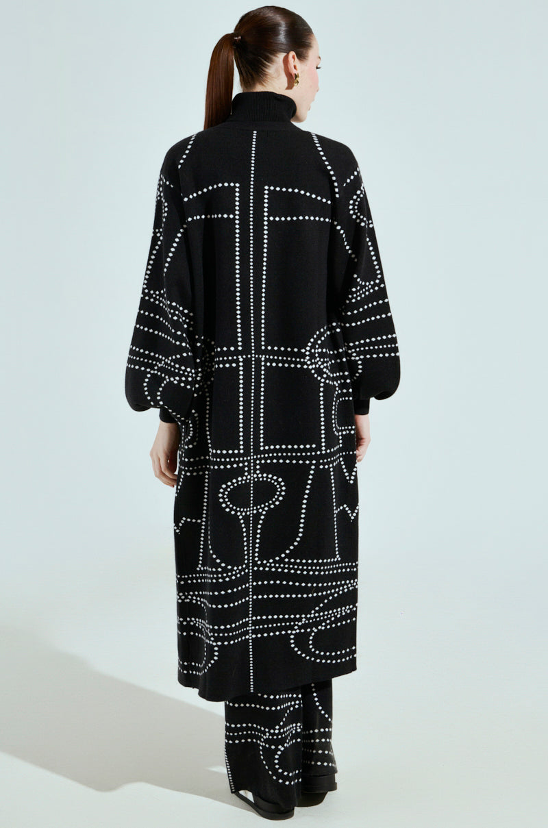 An artsy individual with a ponytail and long brown hair is wearing an oversized black coat and Dotted Oversized Cardigan pants with a white geometric pattern, standing against a light gray background, facing away from the camera.