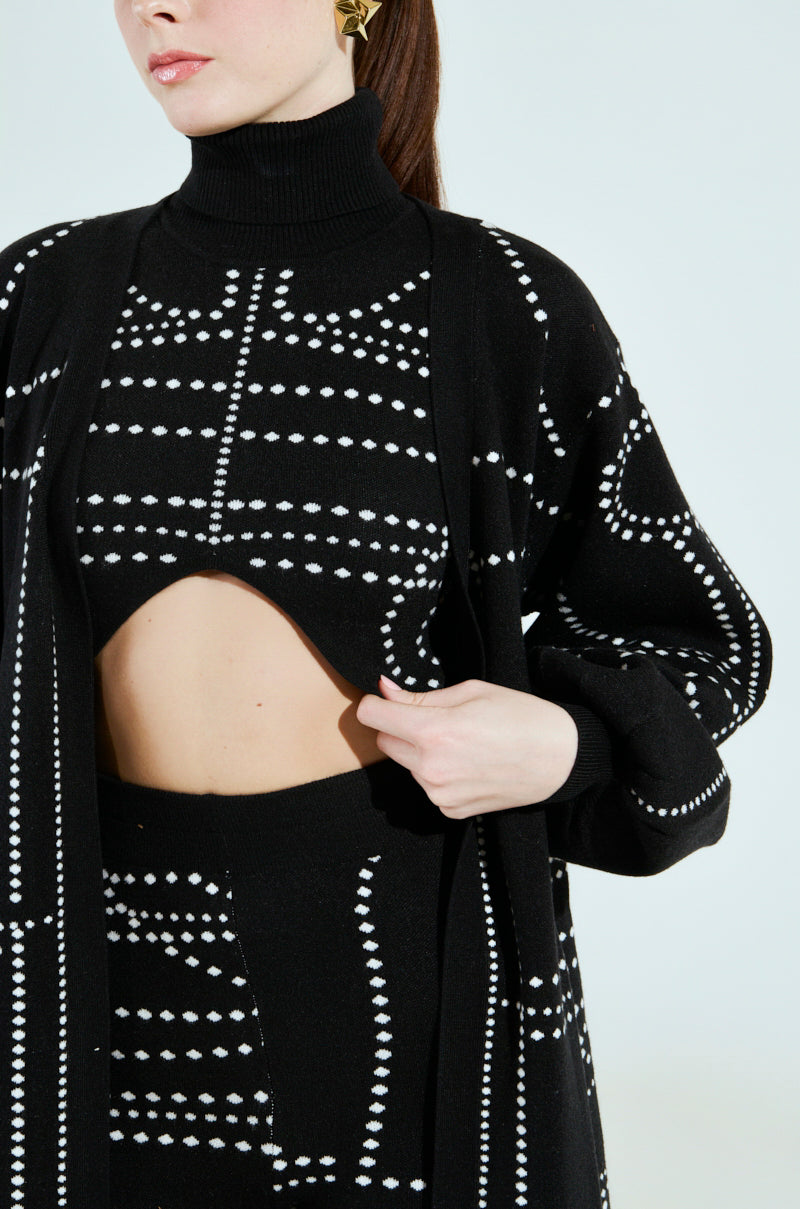 A woman in a DOTTED OVERSIZED CARDIGAN embodies an artsy look with its black and white design, turtleneck, and cut-out midriff. She poses with her hand on the cardigan, complemented by gold geometric earrings that enhance her outfit's abstract pattern.