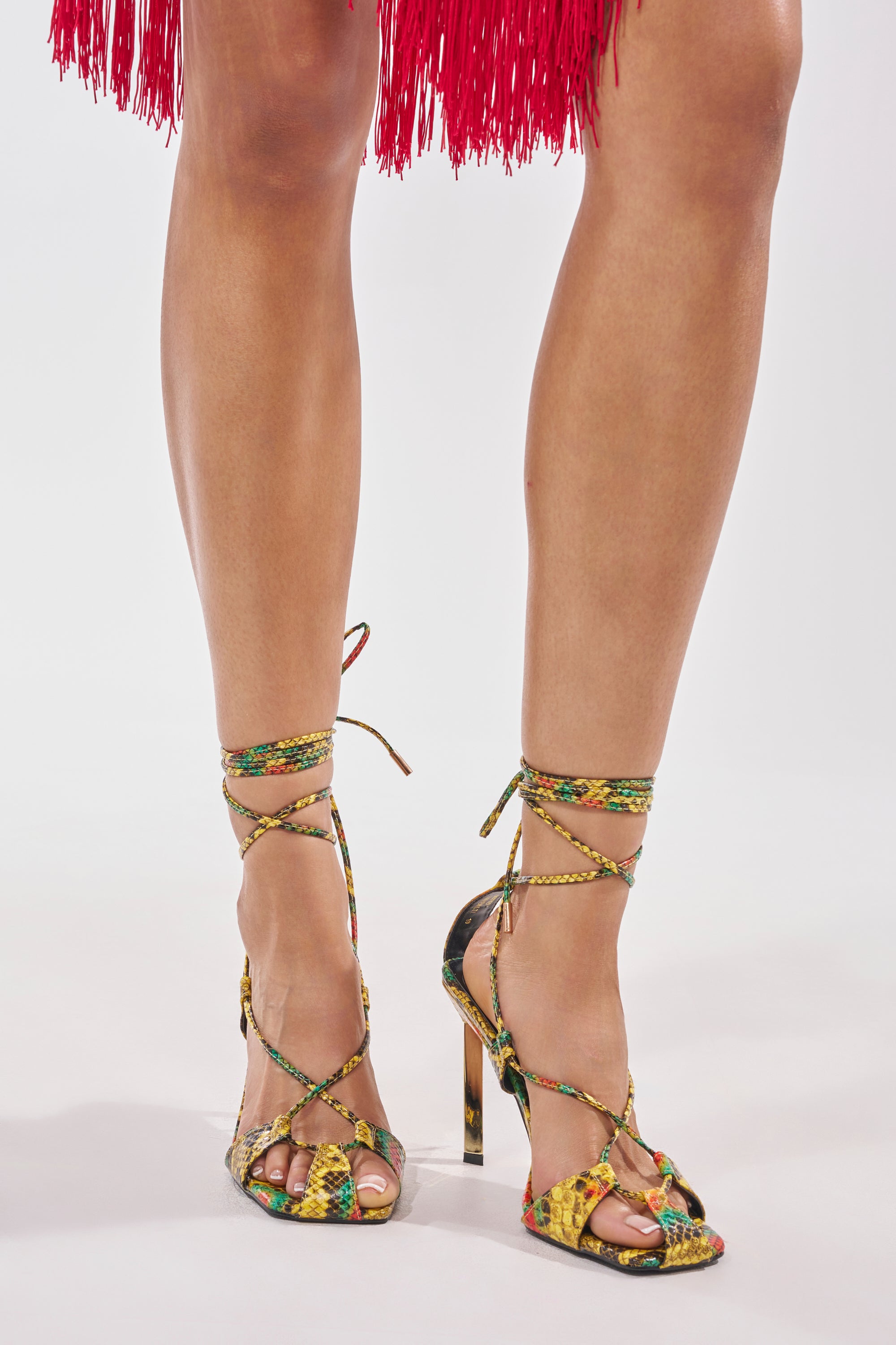 A person stands against a plain white background wearing the AZALEA WANG NOTYET MULTI SNAKE STILETTO SANDAL with colorful lace-up straps, faux leather snakeskin finish, and a red fringed skirt.